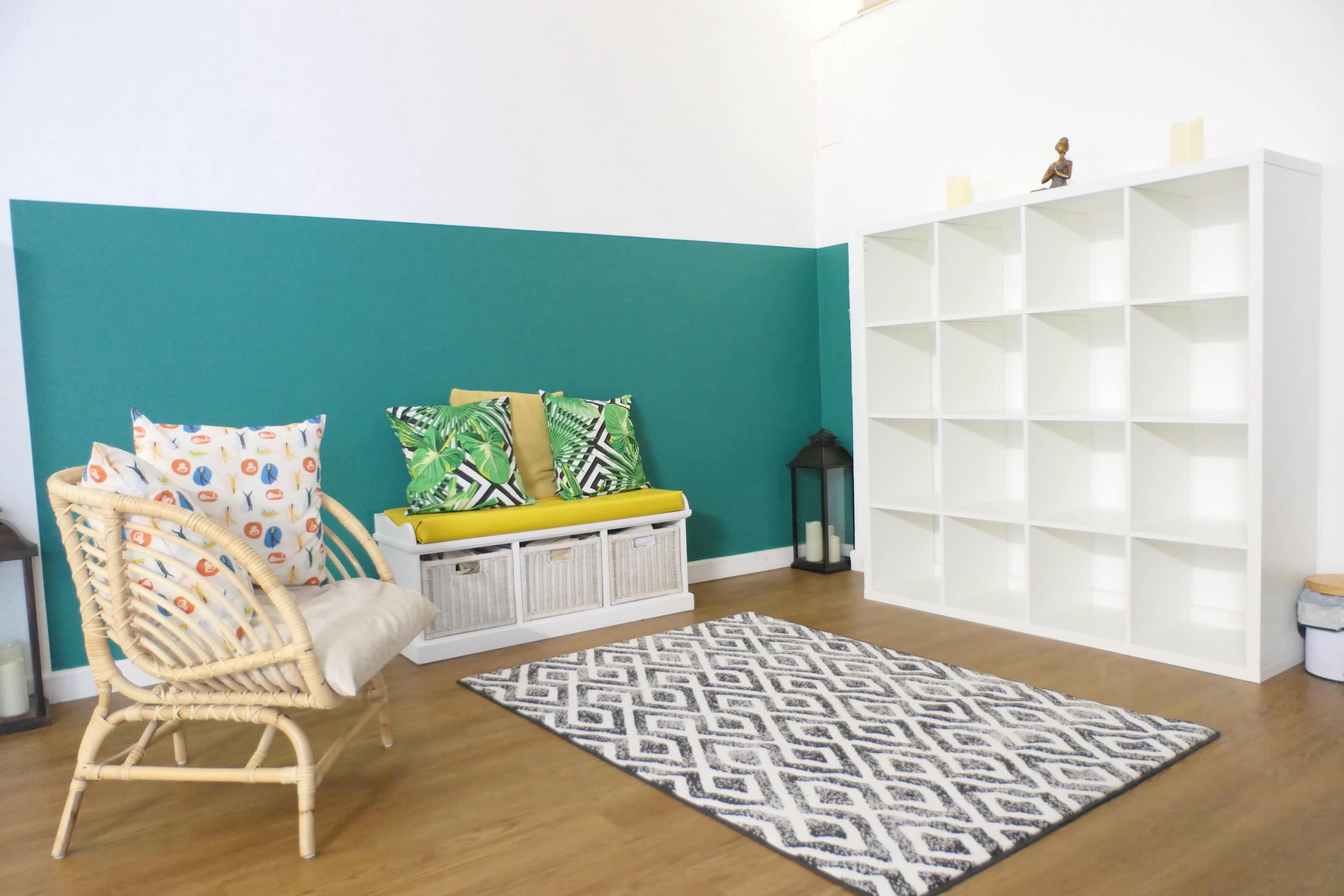 A sitting area featuring a wicker chair, a storage bench with cushions, a patterned rug, and a white shelf against a green accent wall.
