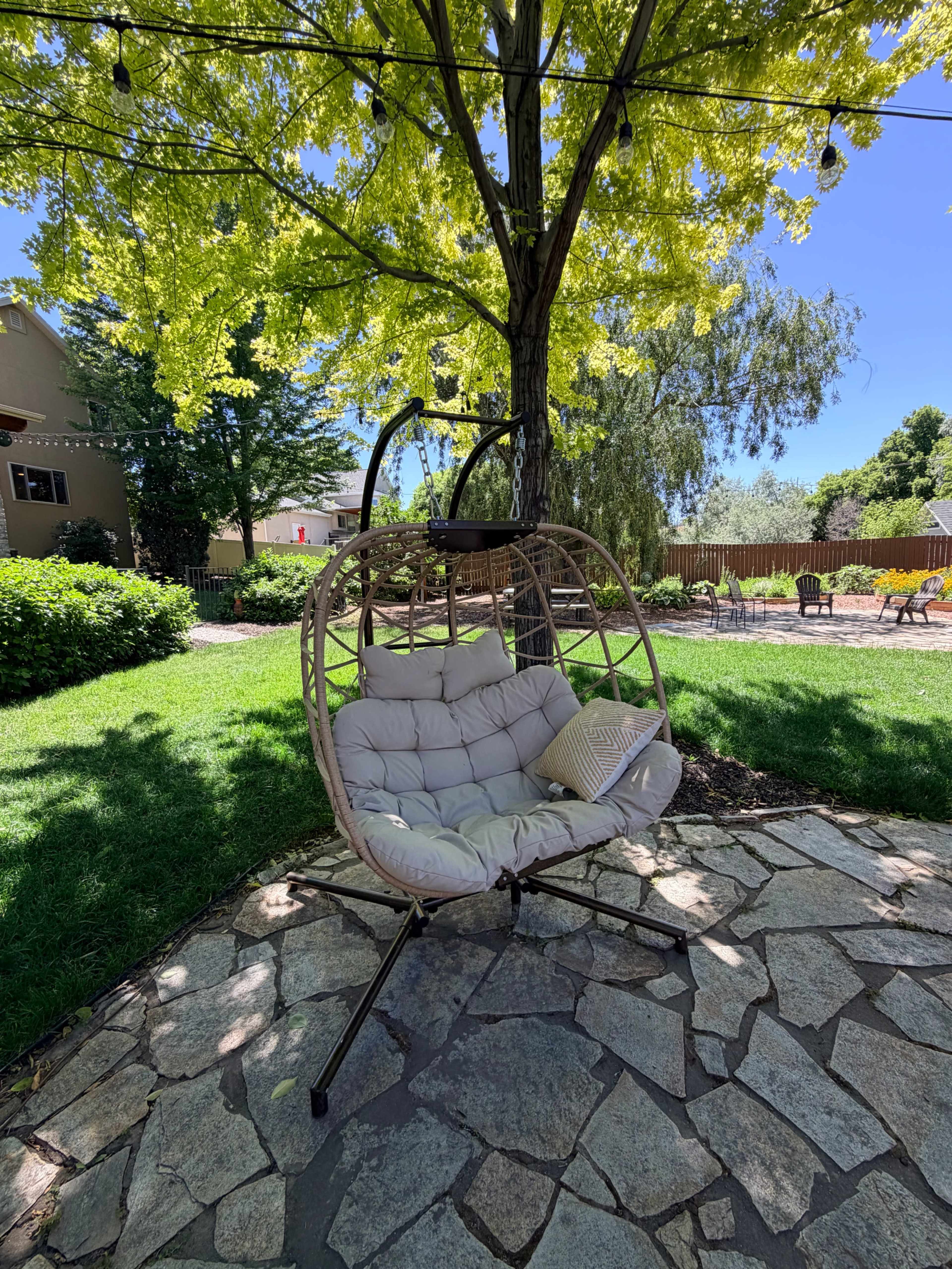 A large, round hanging chair with cushions is positioned on a stone patio beneath a leafy tree in a well-maintained garden.