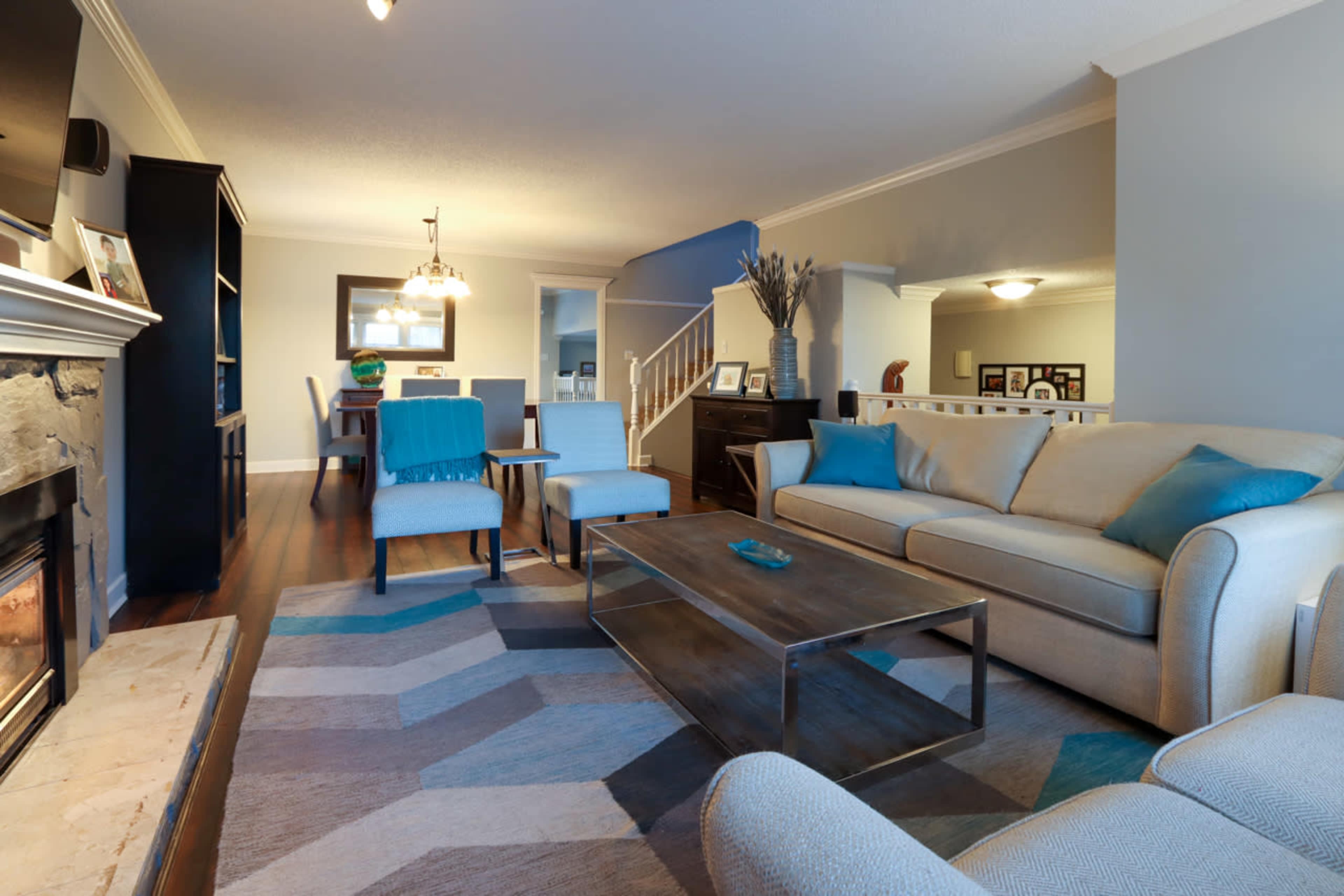 A well-furnished living room features a beige sofa, blue accent pillows, a coffee table, and a fireplace, all arranged on a patterned rug.