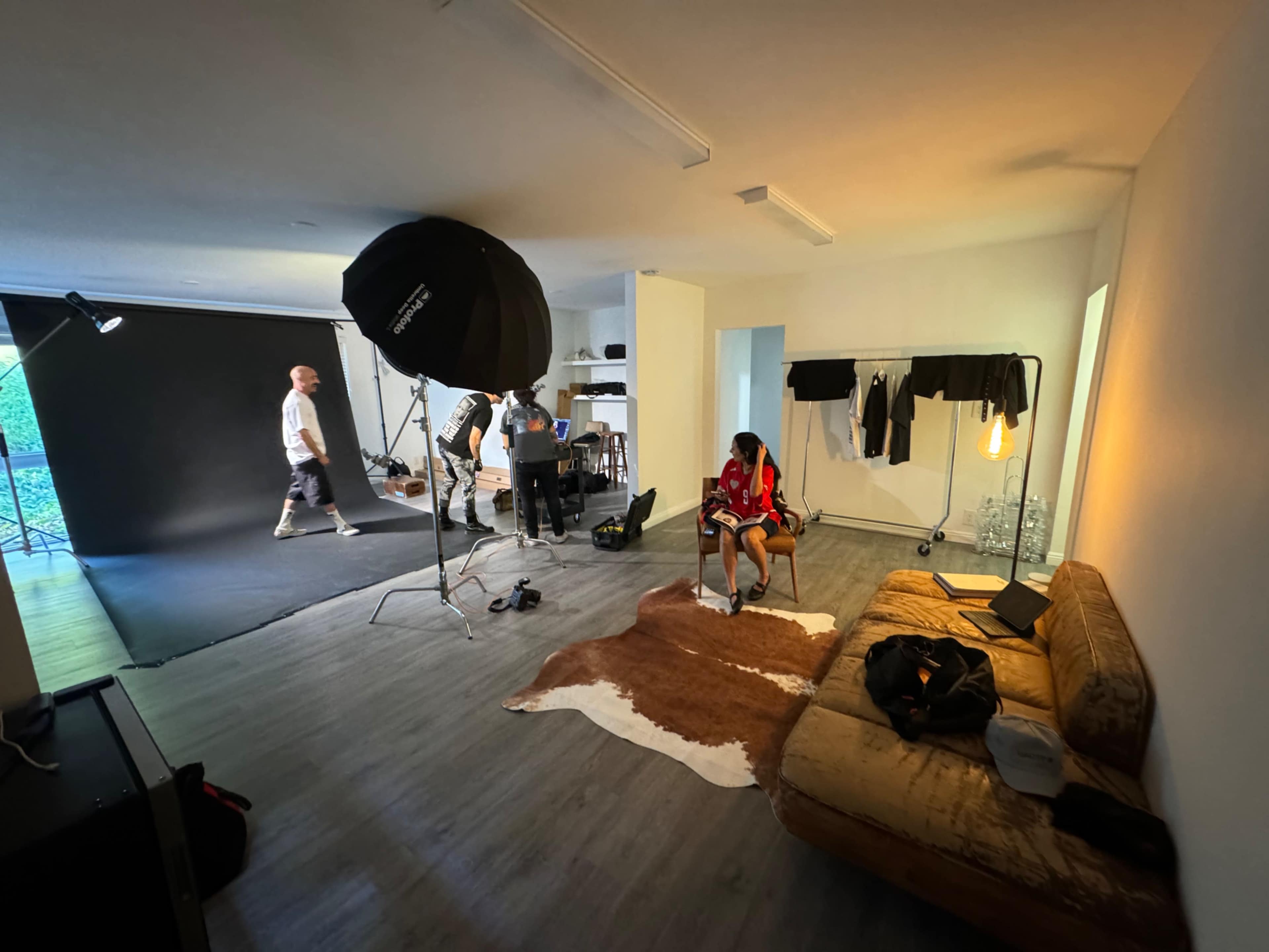 The image shows a photo studio with a model sitting on a couch while a team prepares equipment and backdrops for a photoshoot.