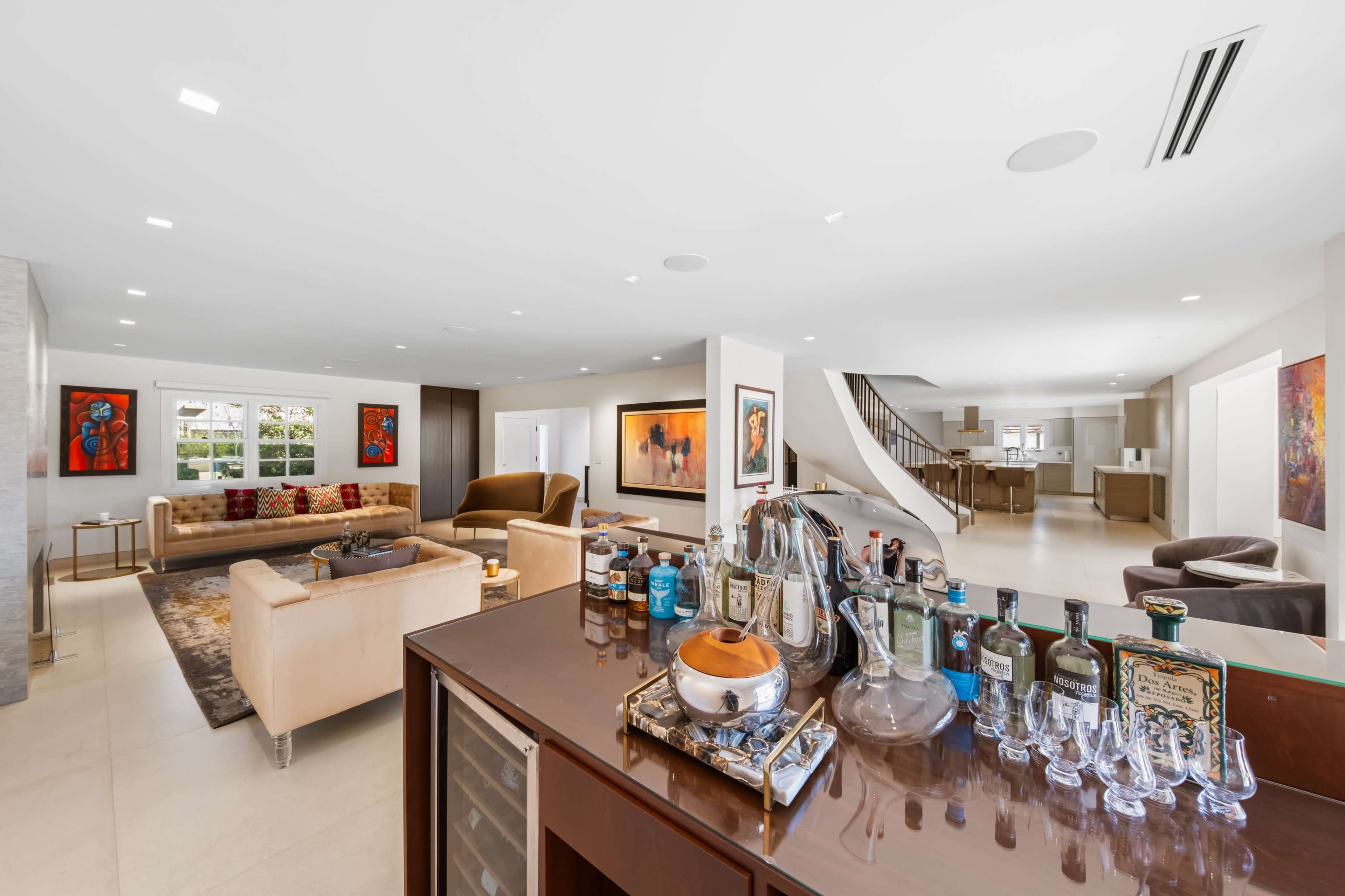 A modern, open-concept living space features a well-stocked bar area with various bottles and glassware, leading into a lounge with sofas and artwork.