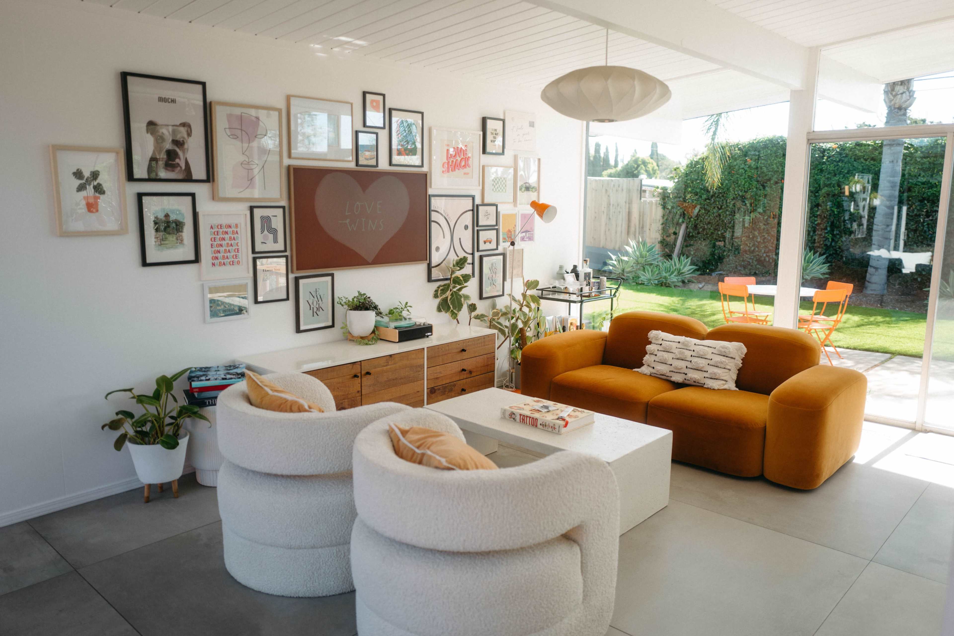 The image shows a bright, modern living room with a mix of artwork on the walls, a cozy orange sofa, two round white chairs, and a wooden coffee table.