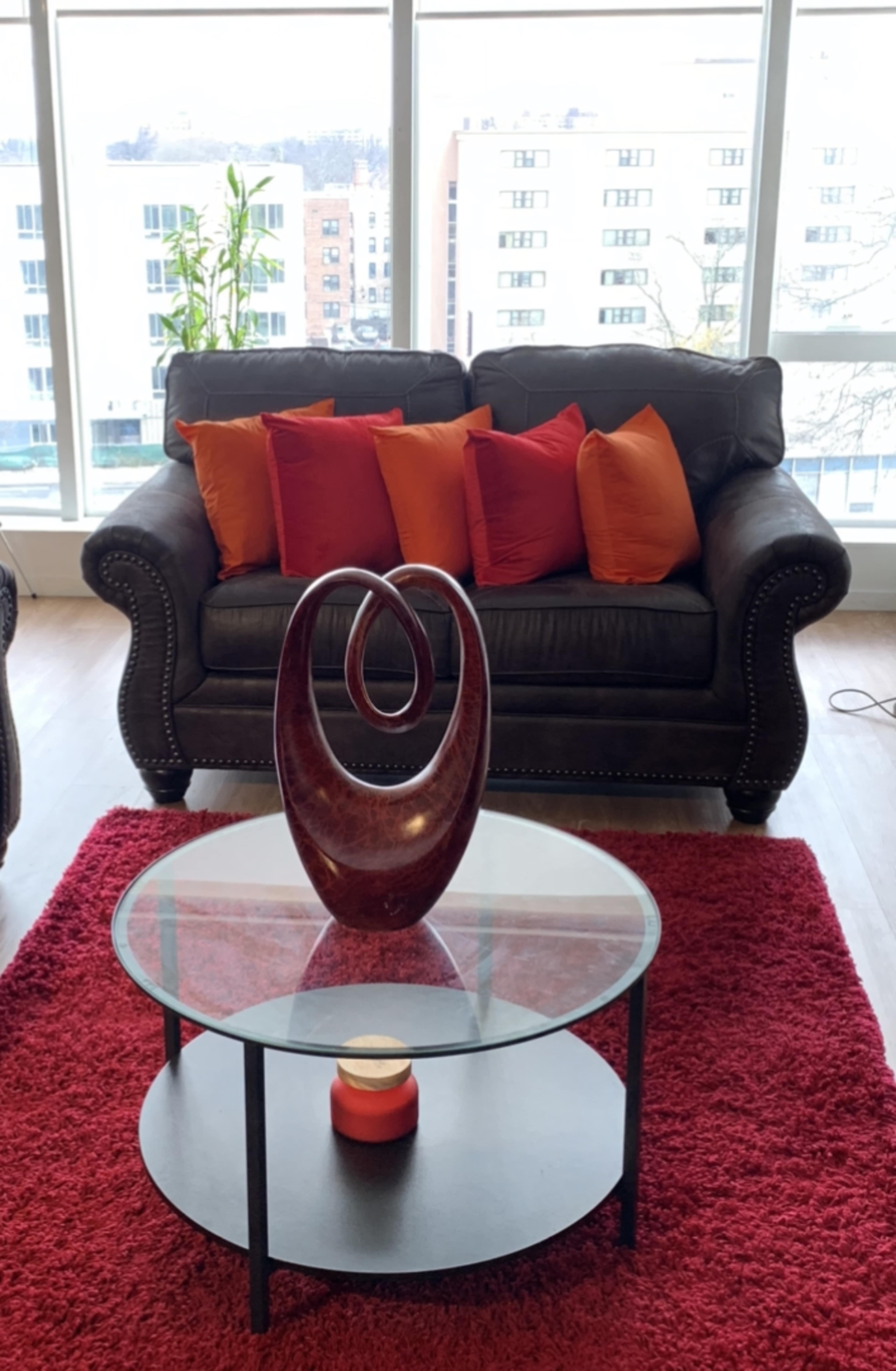 A leather couch with orange and red pillows sits in front of a glass coffee table on a red rug, with a decorative wooden sculpture and a candle on the table.