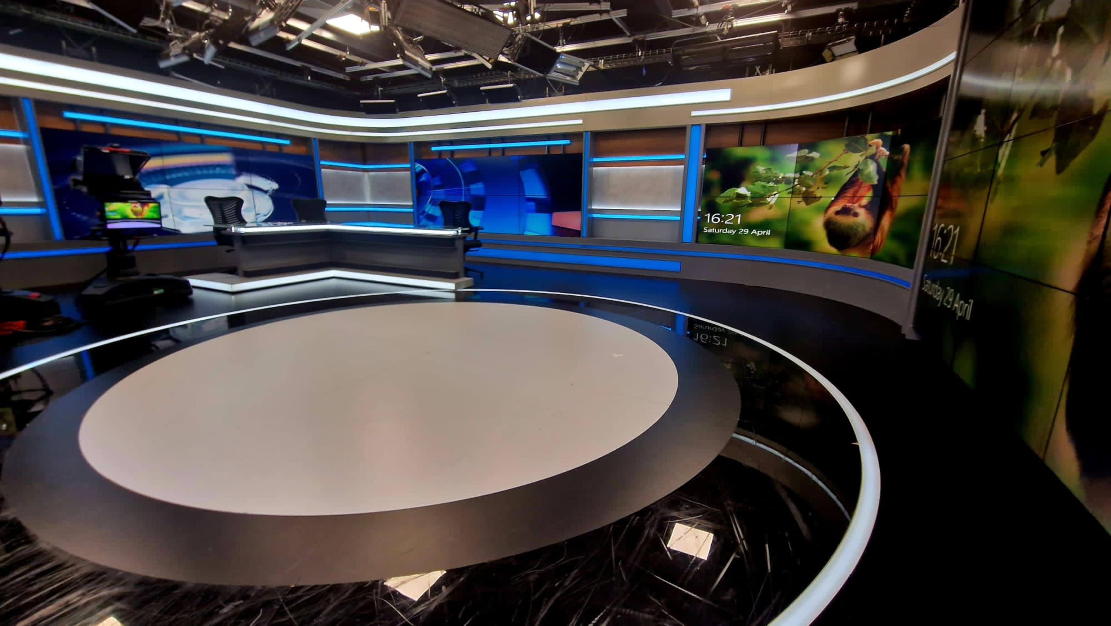 The image shows a modern news studio with a circular stage area, large screens displaying information, and a backdrop featuring a nature scene.