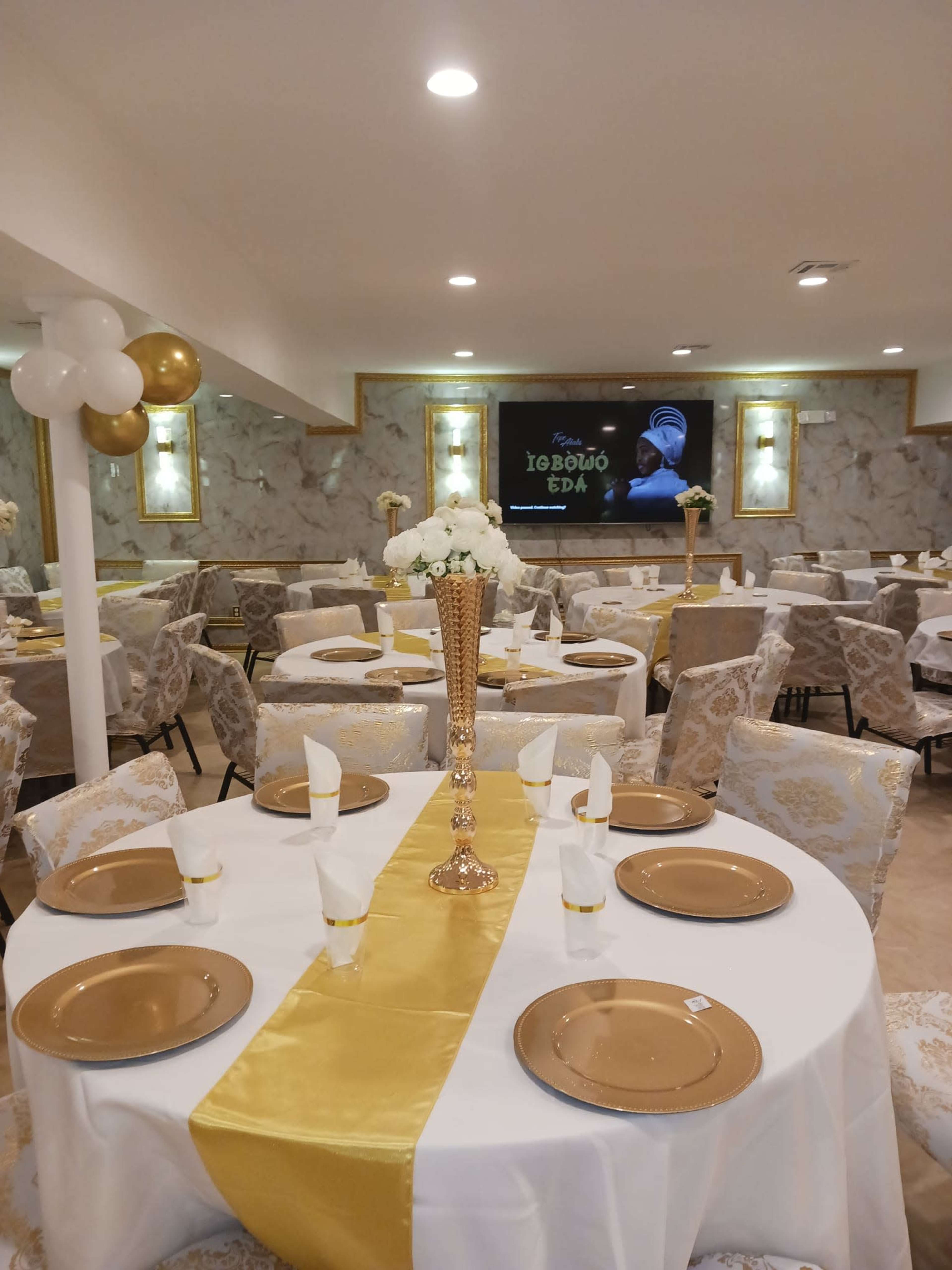 A banquet hall set up with elegantly decorated tables featuring gold accents, centered around a large screen displaying a video.