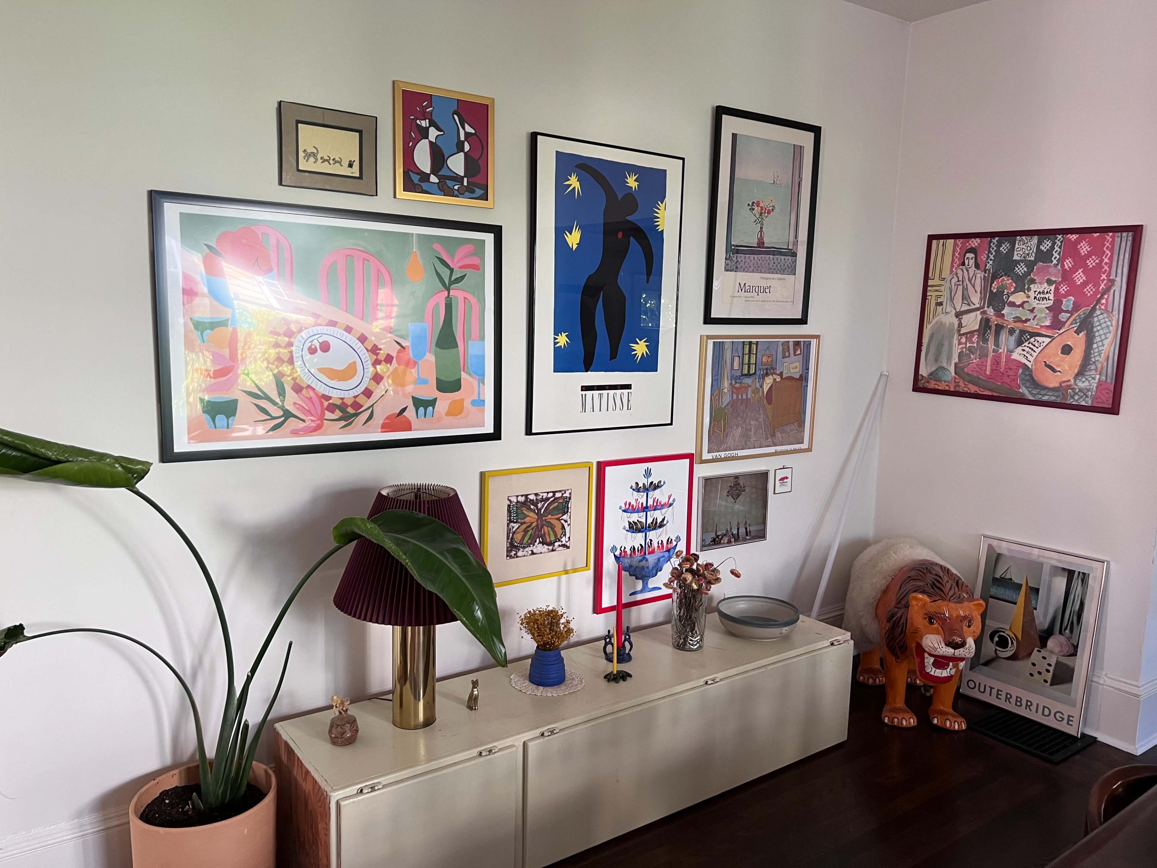 A gallery wall adorned with various framed artworks, alongside a potted plant and a decorative side table in a well-lit room.