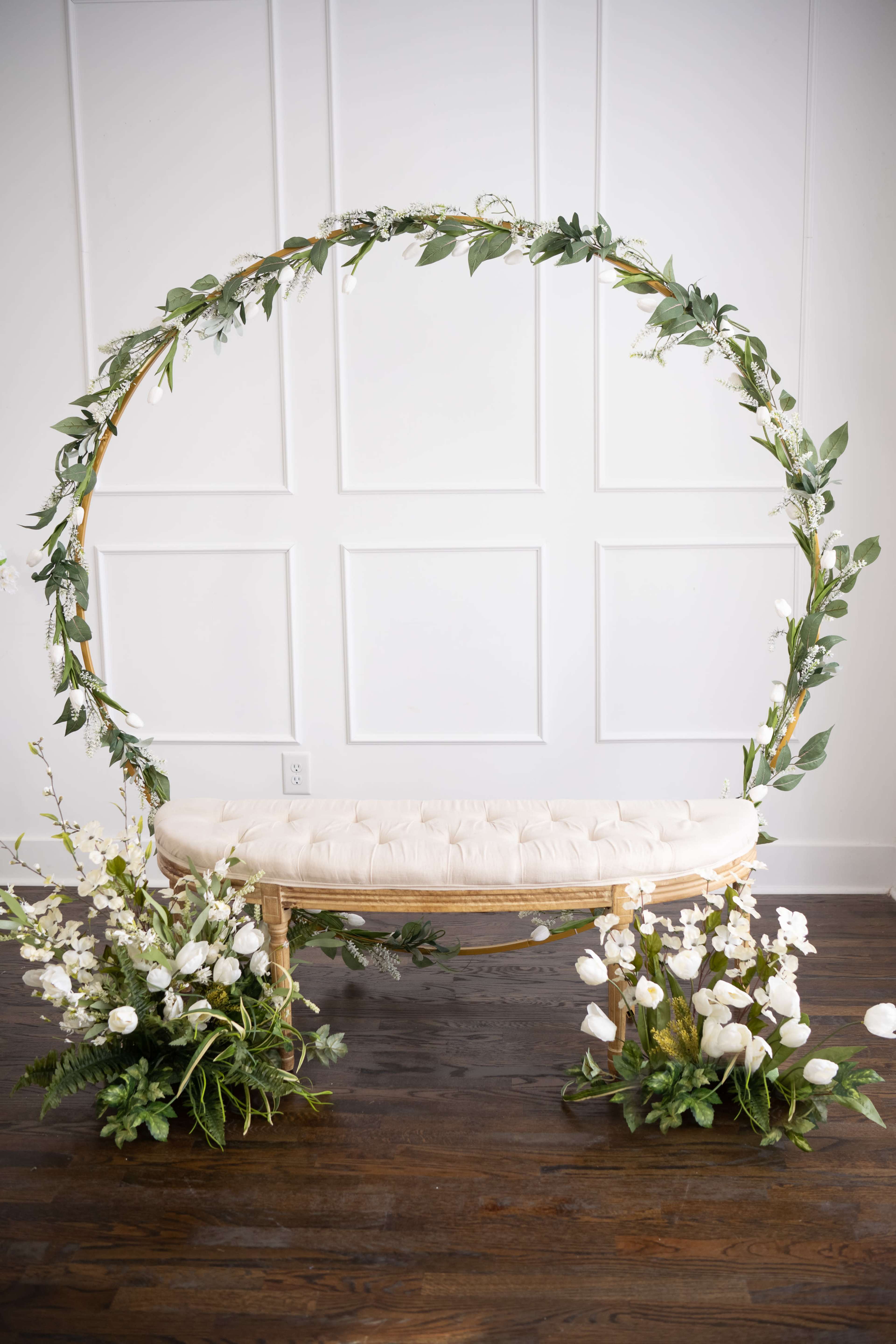 An elliptical floral arch surrounds a tufted bench adorned with white flowers and green foliage on each side, set against a white paneled wall.