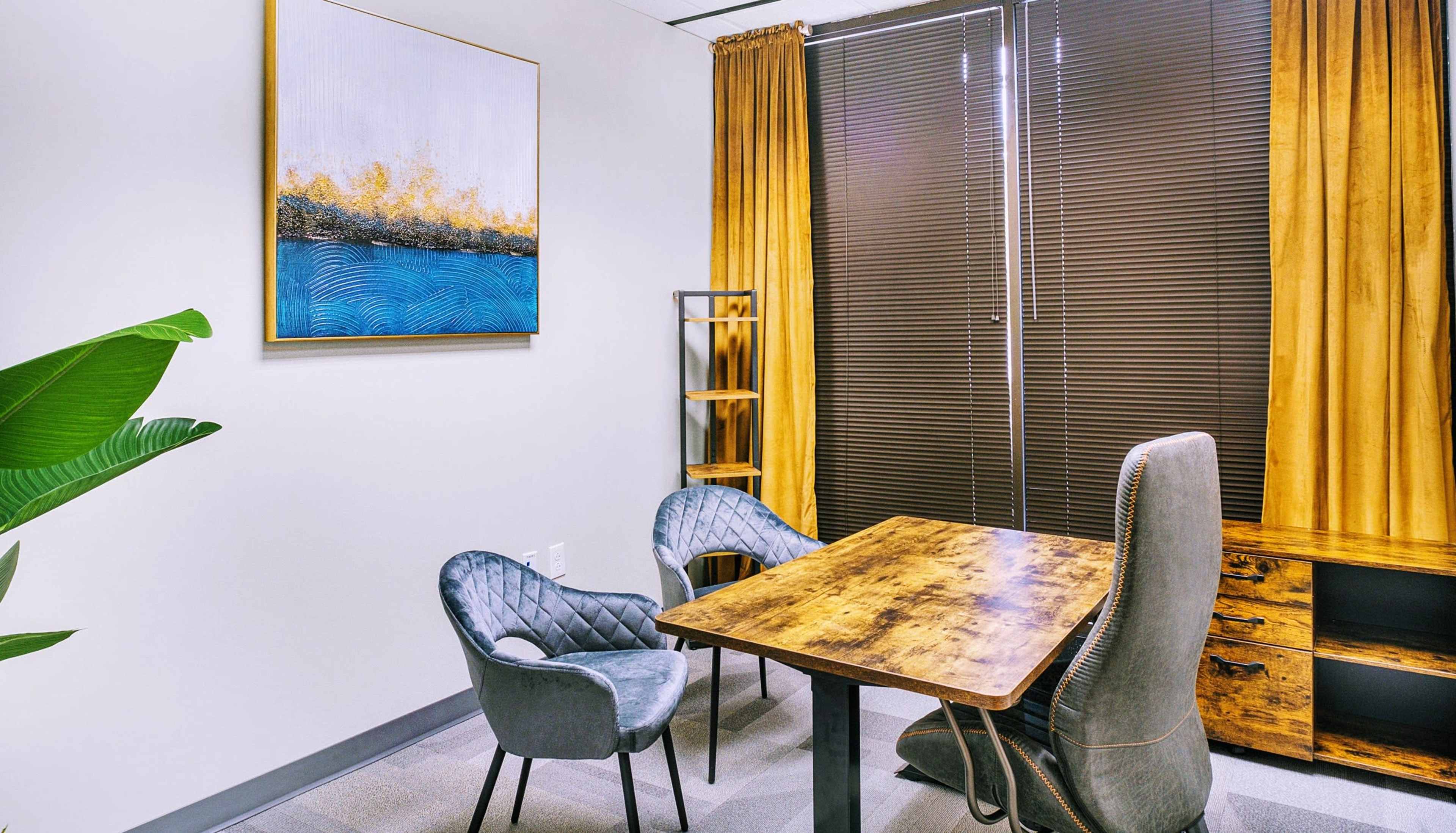 A small office space featuring a wooden table, two gray chairs, and a large abstract painting on the wall, with floor-to-ceiling windows covered by dark blinds.
