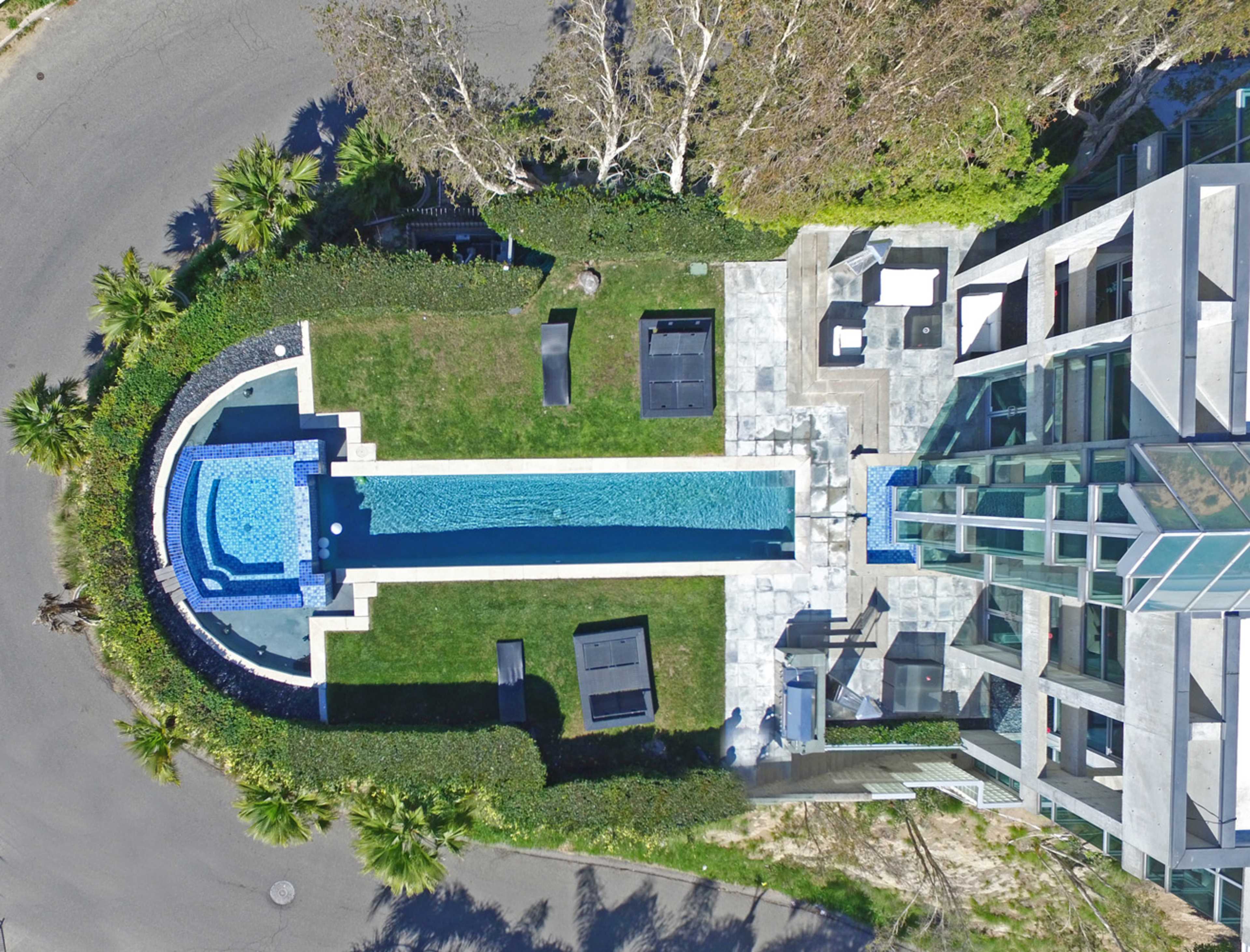 An aerial view shows a modern, rectangular swimming pool surrounded by landscaped greenery and lounge chairs next to a contemporary building.