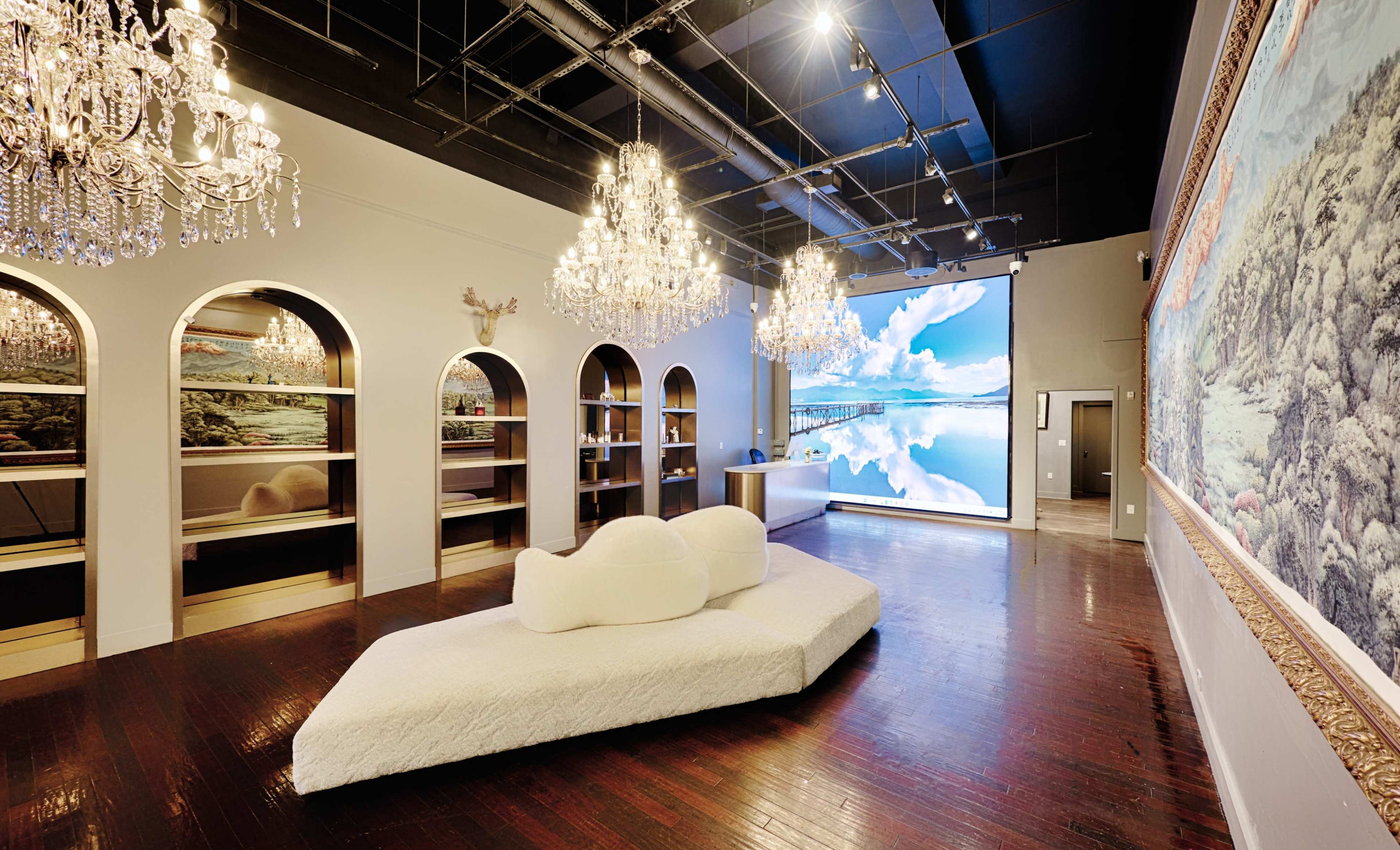 The image shows a spacious interior with chandeliers, a large digital display, and a minimalist white couch in a well-lit gallery setting.