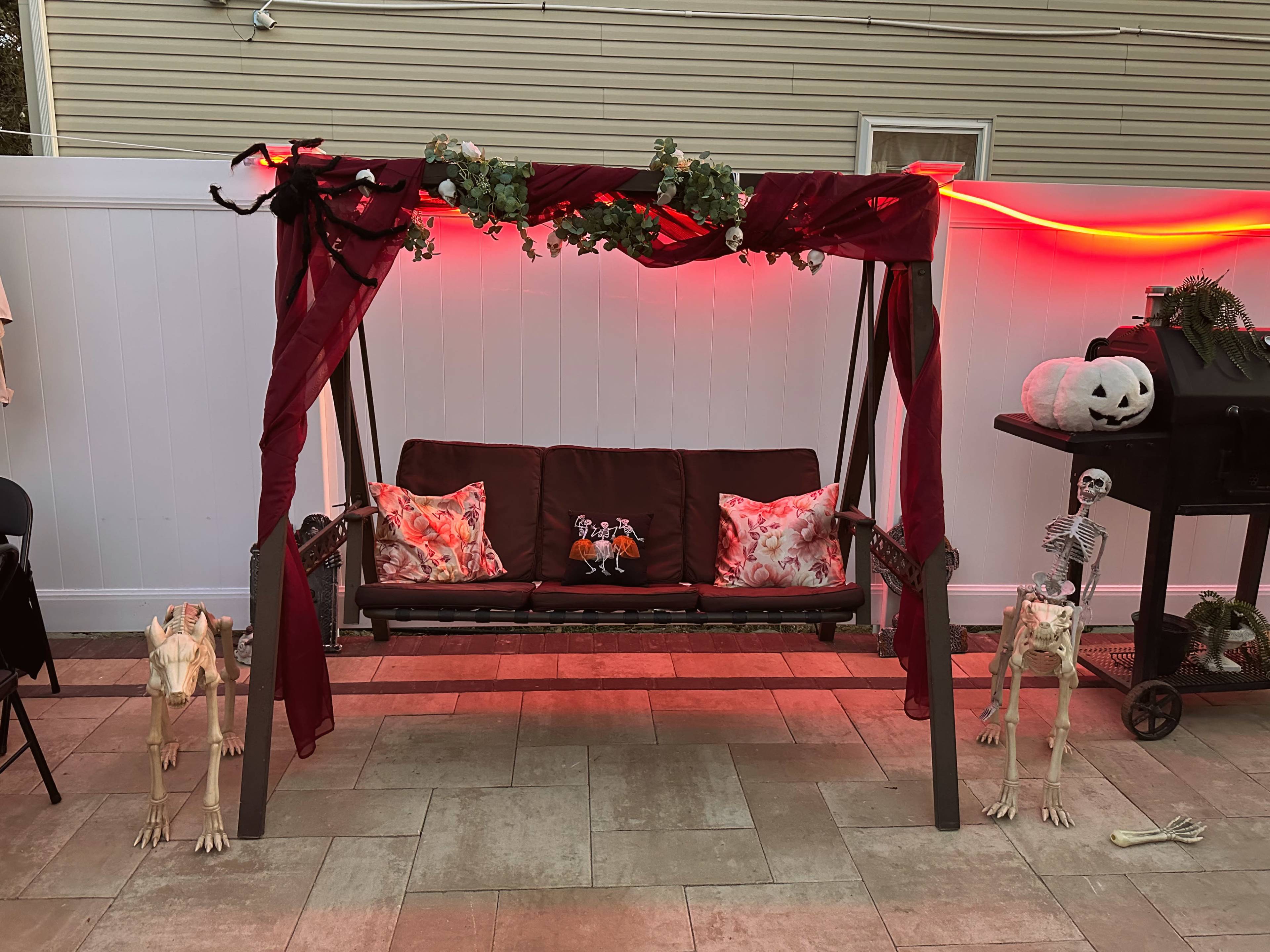 A porch swing is draped with red fabric and decorated with faux vines, flanked by skeleton figures, amidst a festive setting.