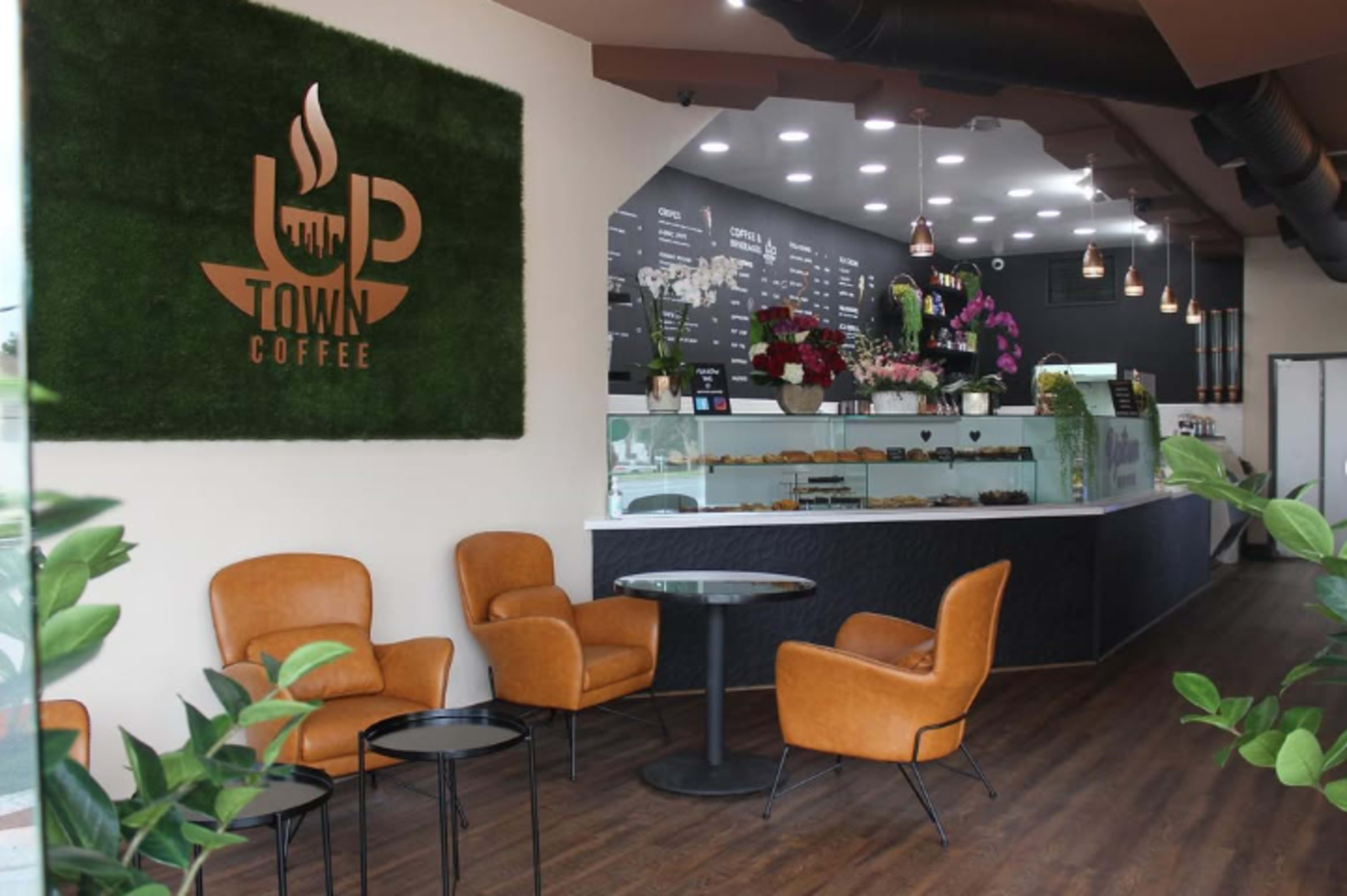 The image shows a modern coffee shop interior featuring brown leather chairs, a circular table, and a counter with a display of baked goods, adorned with plants and decorative flowers.