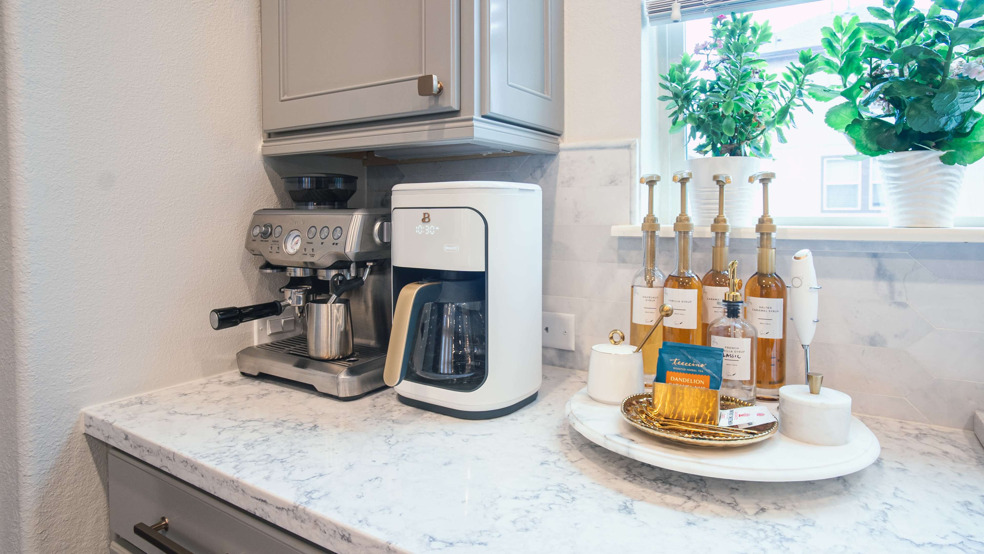 The image shows a kitchen countertop with a coffee machine, a drip coffee maker, and a set of elegant bottles on a round marble tray, next to a window with potted plants.