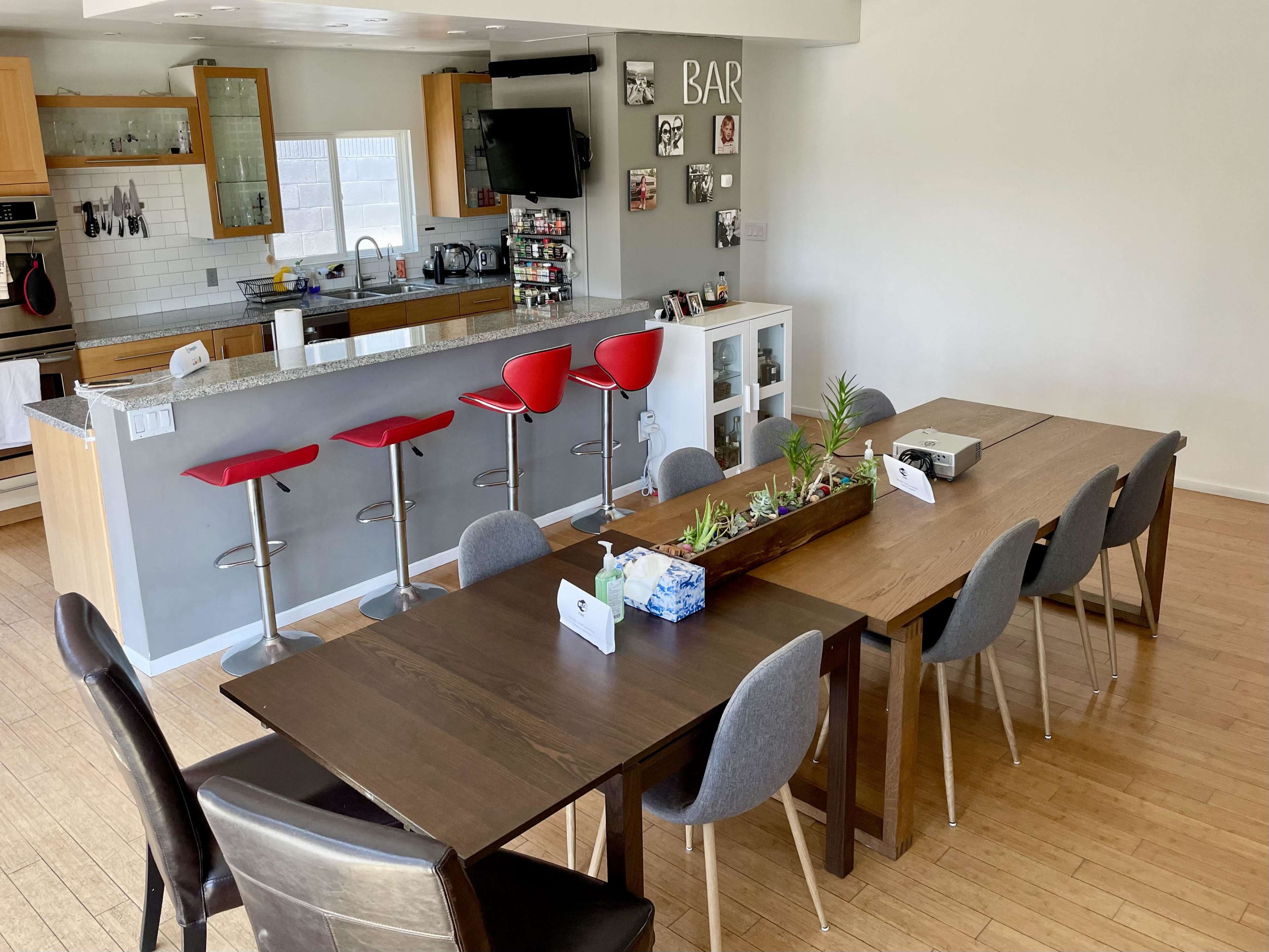 A spacious dining area features a long wooden table surrounded by chairs, with a bar area in the background lined with red stools.