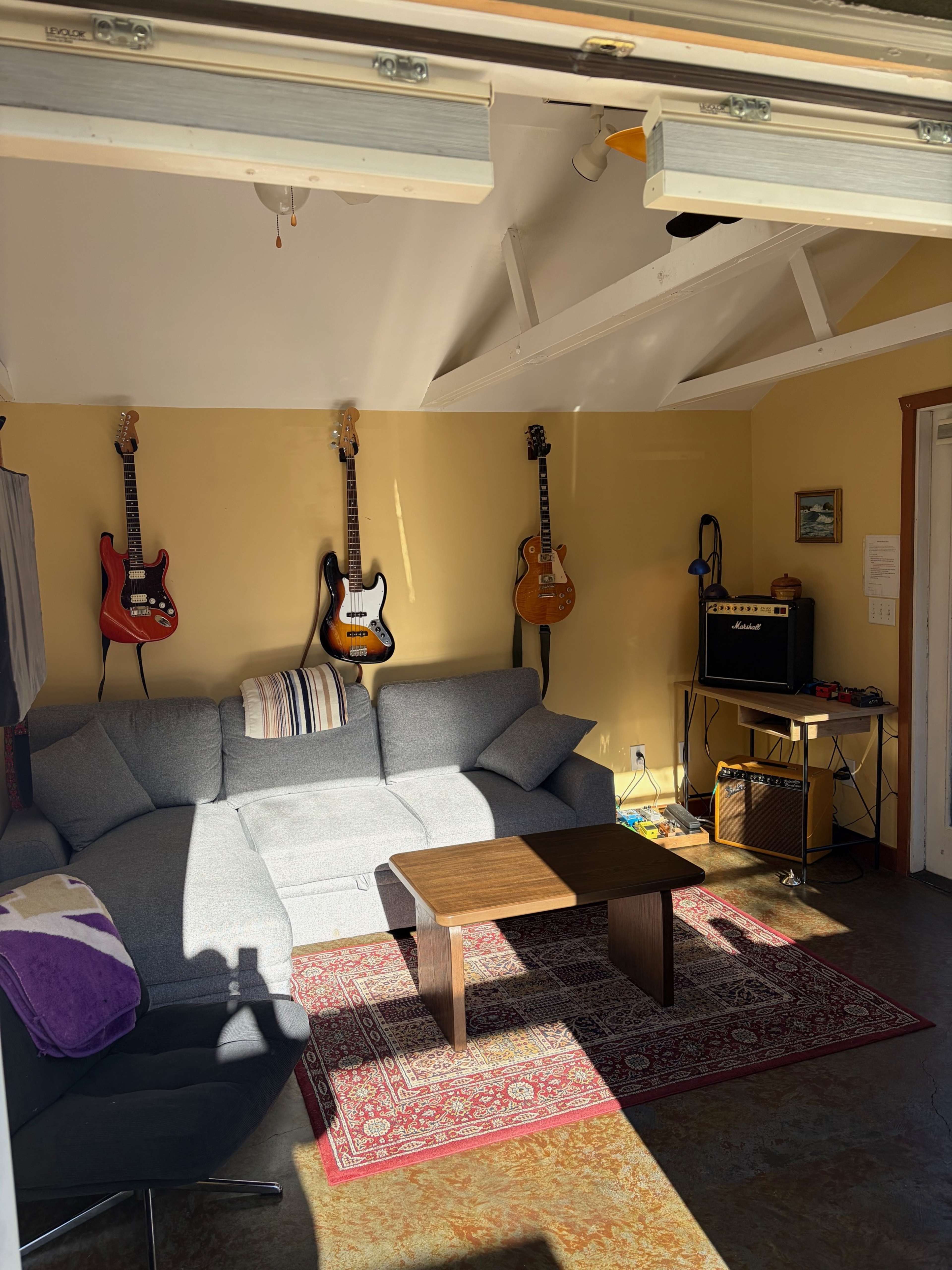The image shows a cozy room with a sectional sofa, a wooden coffee table, and several guitars mounted on the wall, illuminated by natural light from a garage door.