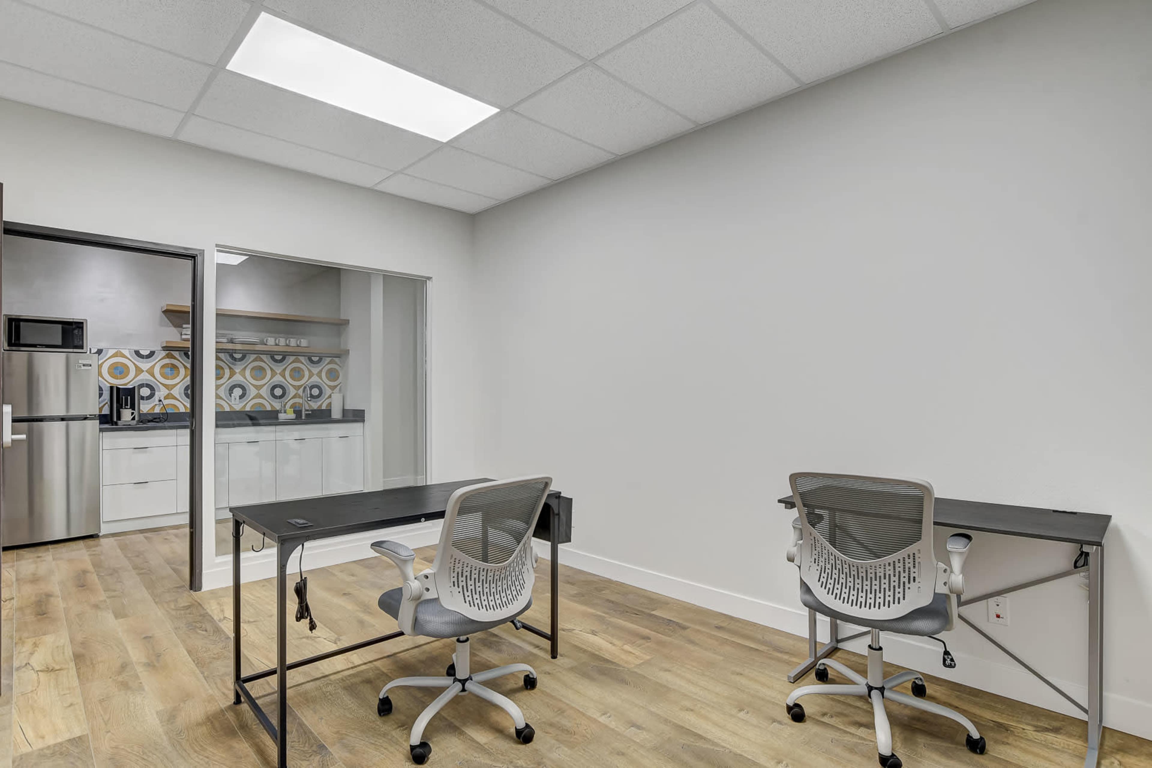 A small office space with two desks and chairs, a kitchen area visible in the background, and a light-colored wall.