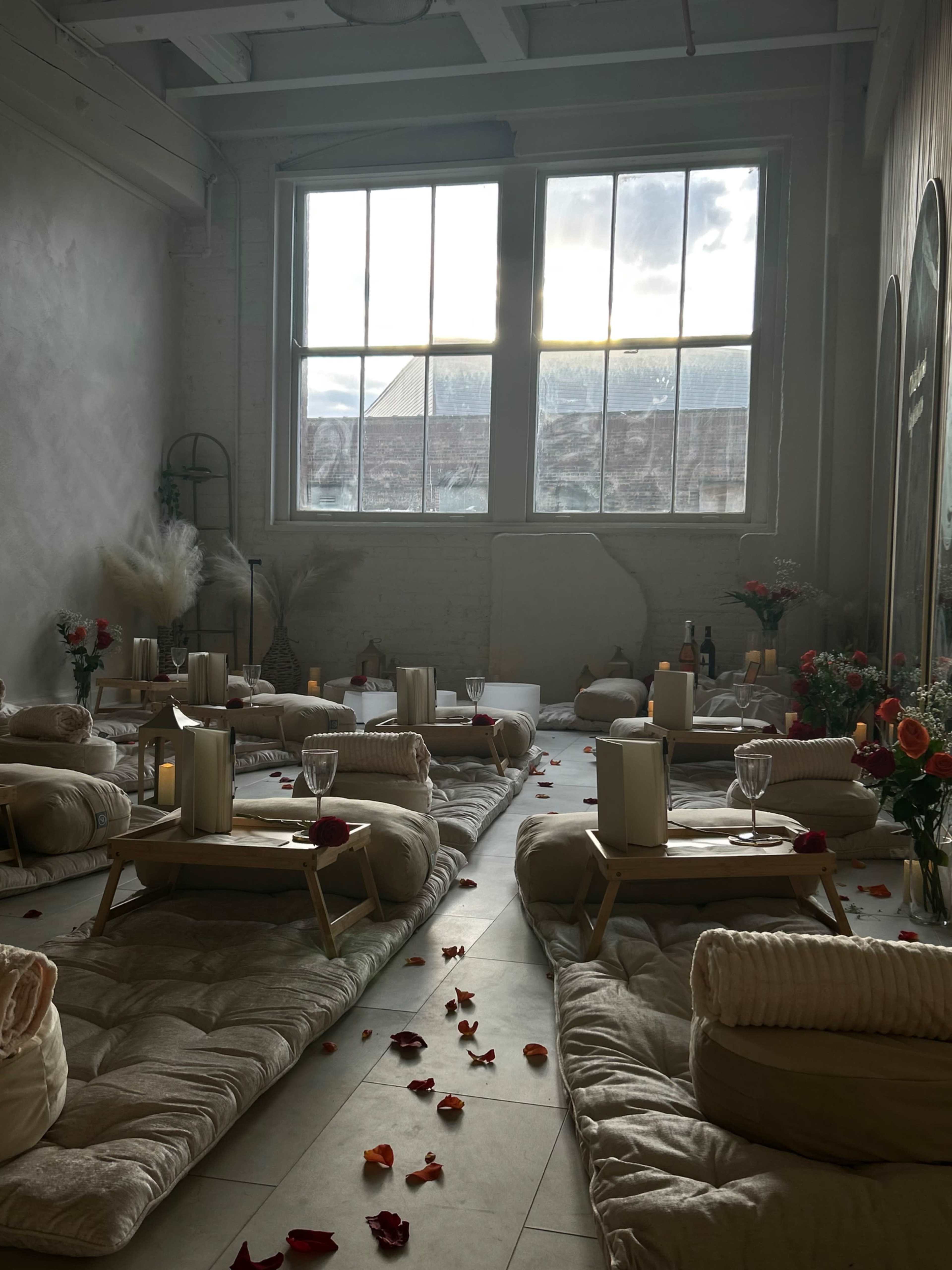 The image shows a minimalistic room with several low seating areas on mats, adorned with flower petals and candles, illuminated by sunlight filtering through large windows.