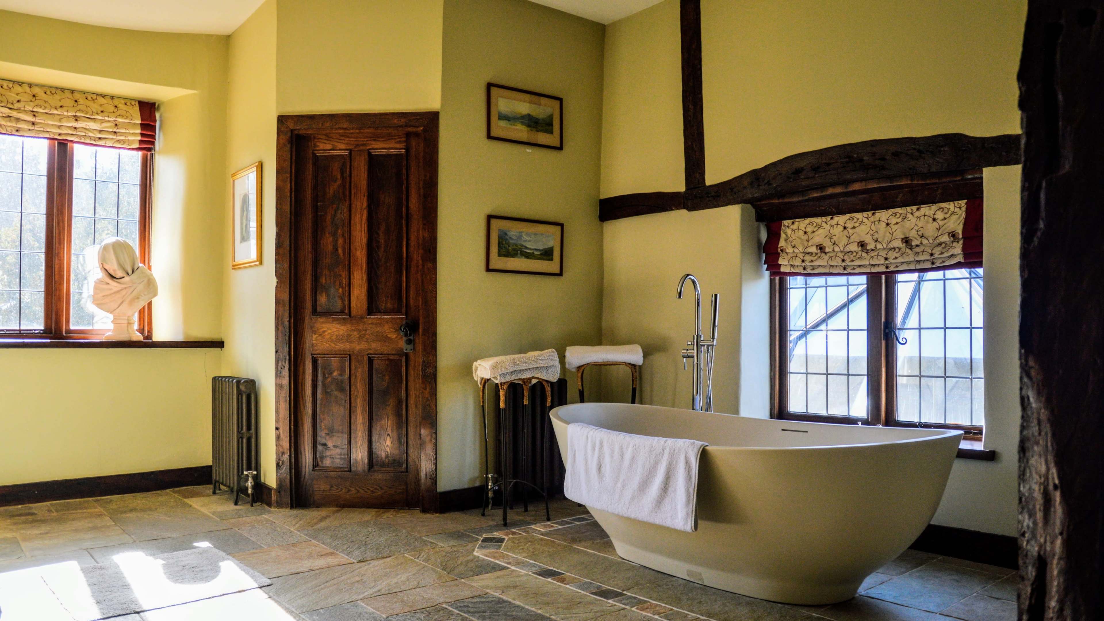 A bright bathroom featuring a freestanding bathtub, wooden door, and a window with a person draped in a towel.