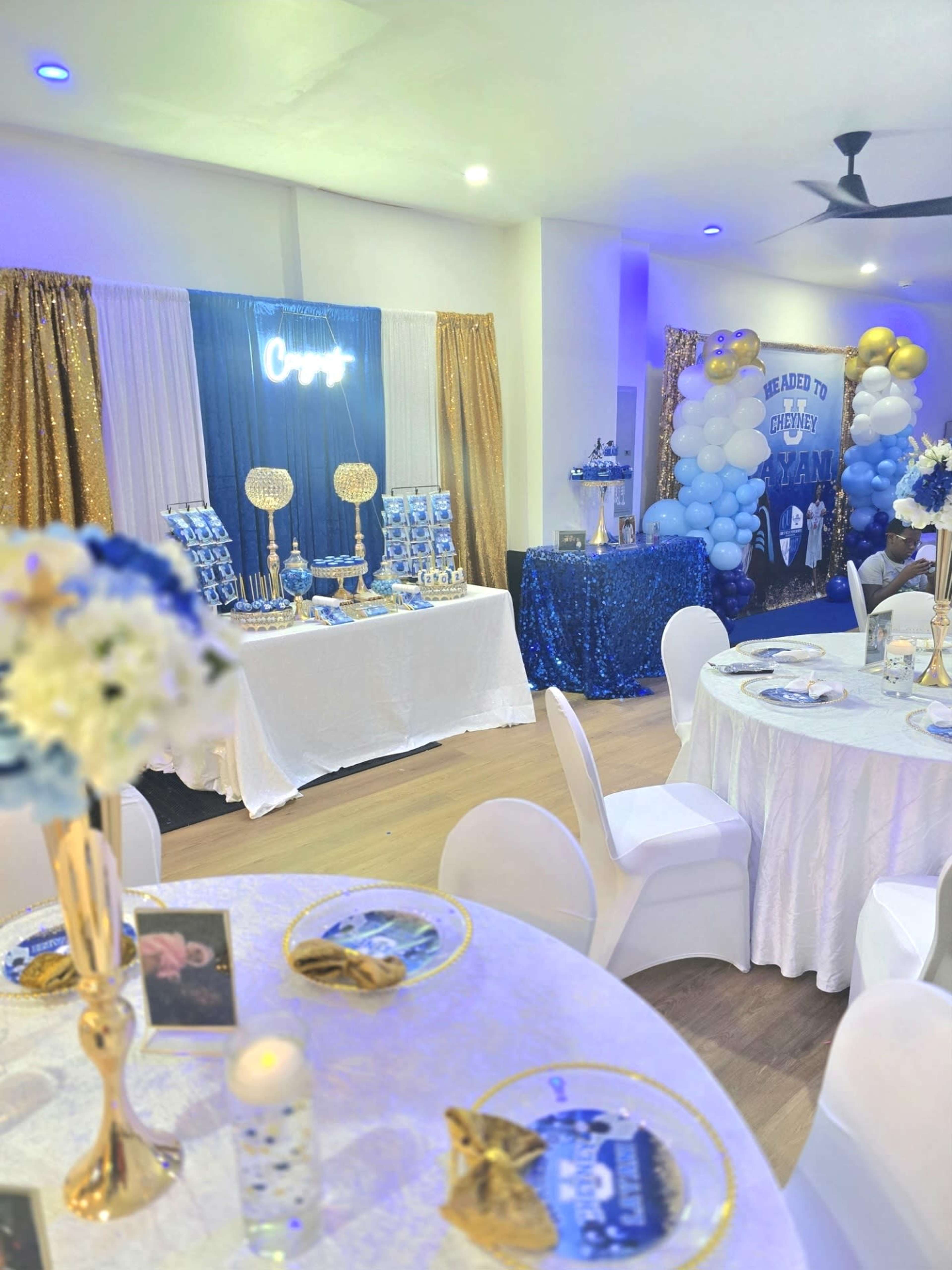The image shows a decorated event space featuring tables with white tablecloths, a dessert table with blue and gold accents, and a backdrop adorned with balloons.