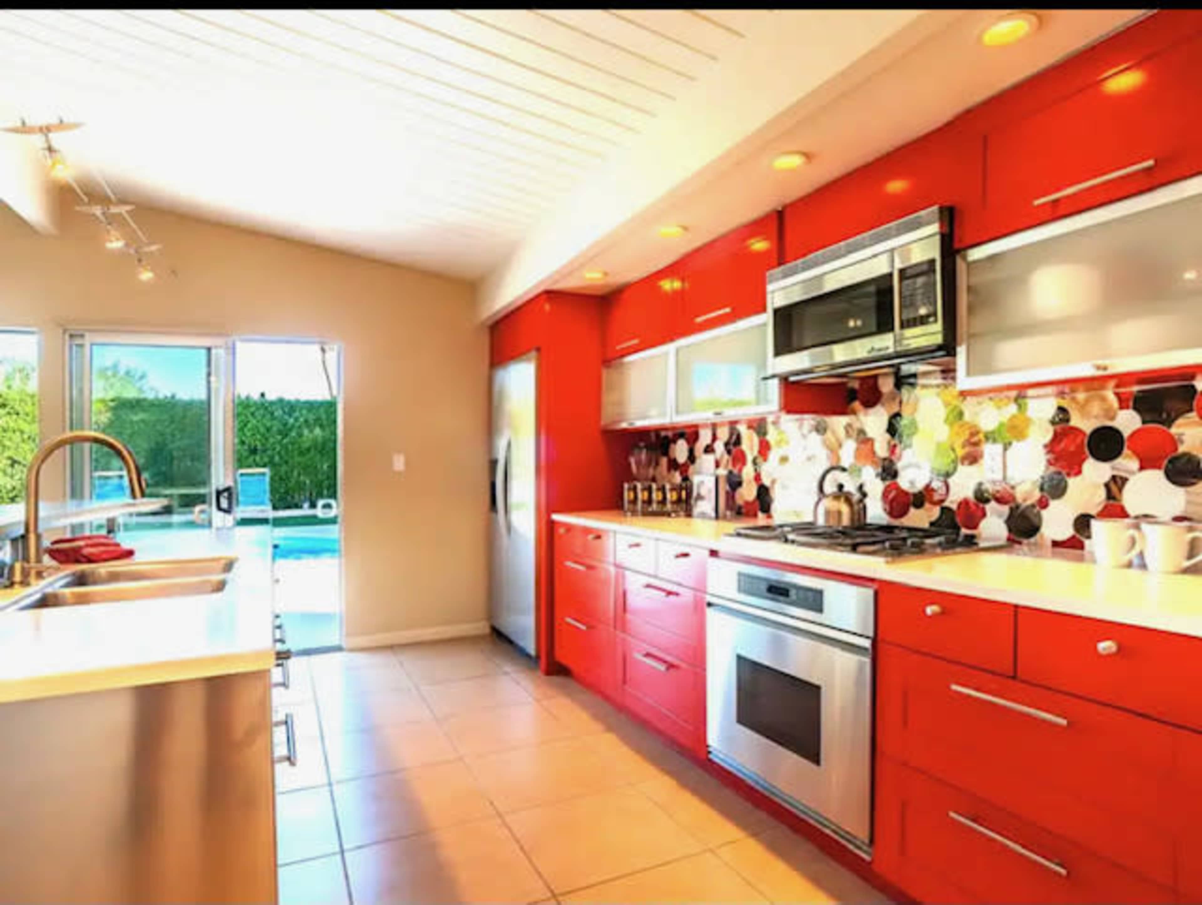 The modern kitchen features bright red cabinetry, stainless steel appliances, and a tile backsplash with circular patterns, overlooking a pool area through sliding glass doors.
