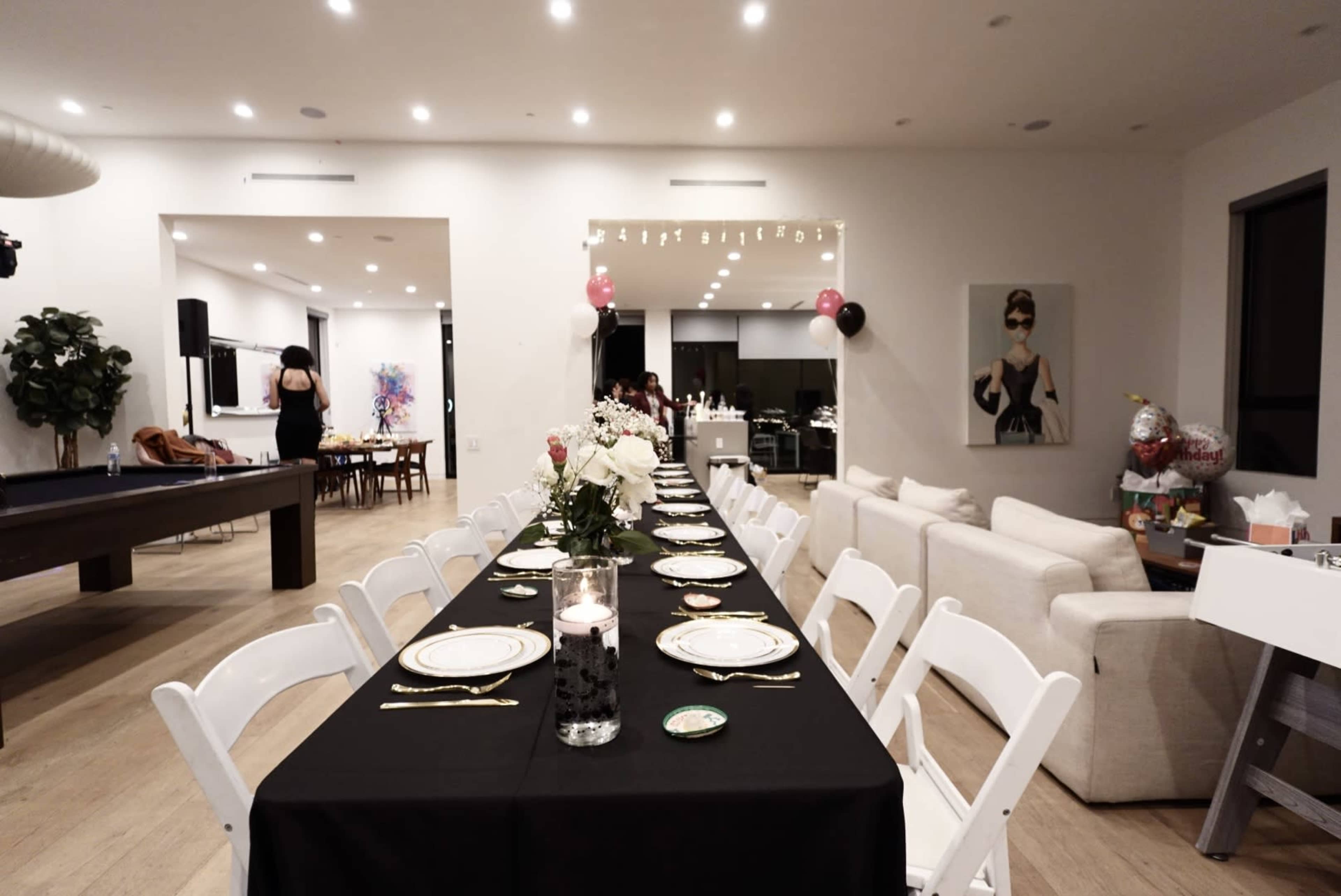 A long black table is set for a dinner party in a modern dining area, with white chairs and decor including flowers and candles, alongside a pool table and an open kitchen.