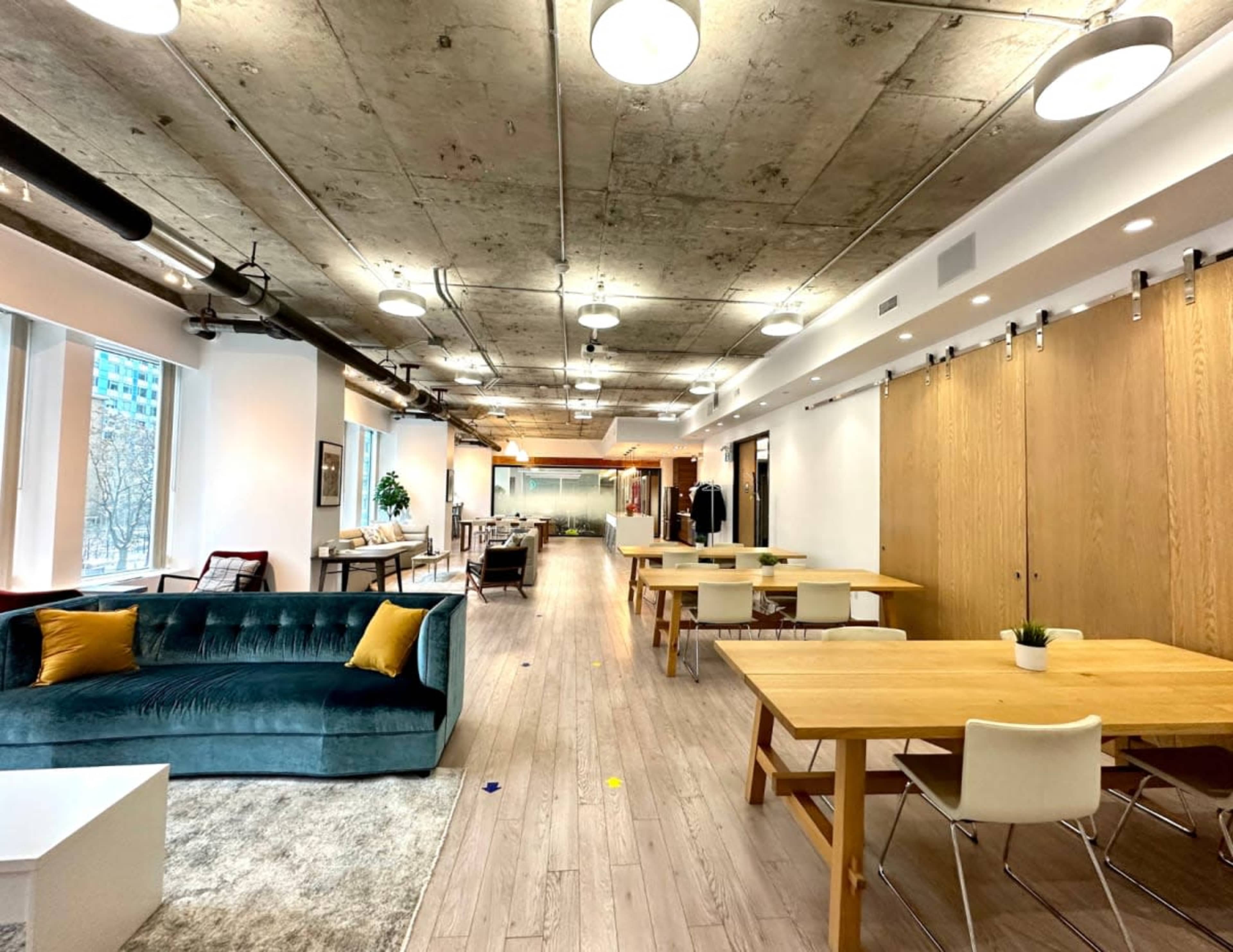 The image shows a spacious office interior with modern furniture, including a green sofa, wooden tables, and a textured ceiling.
