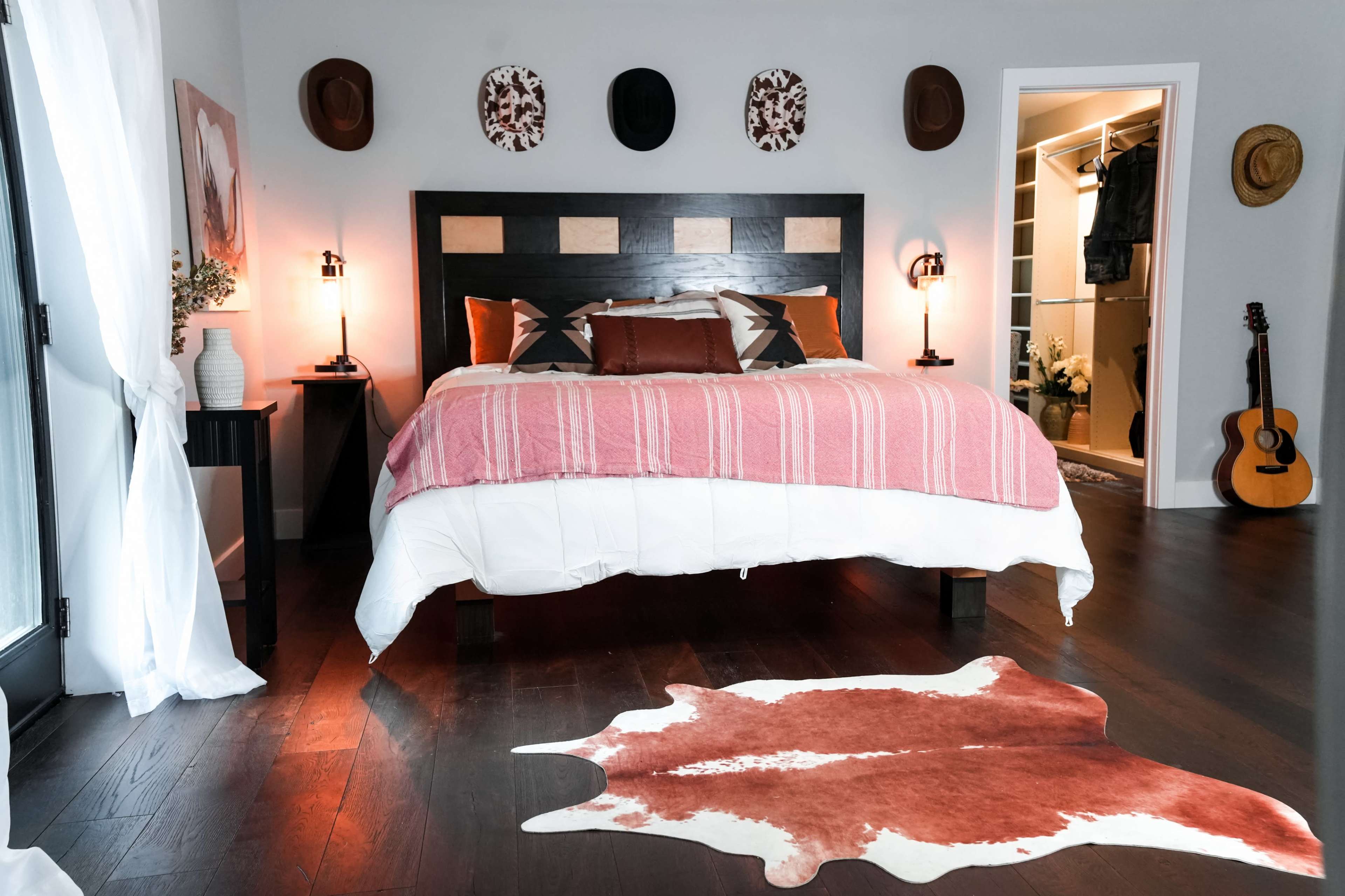 The image depicts a stylish bedroom featuring a large bed with a pink-striped throw blanket, a cowhide rug on the dark wood floor, and a guitar leaning against the wall.