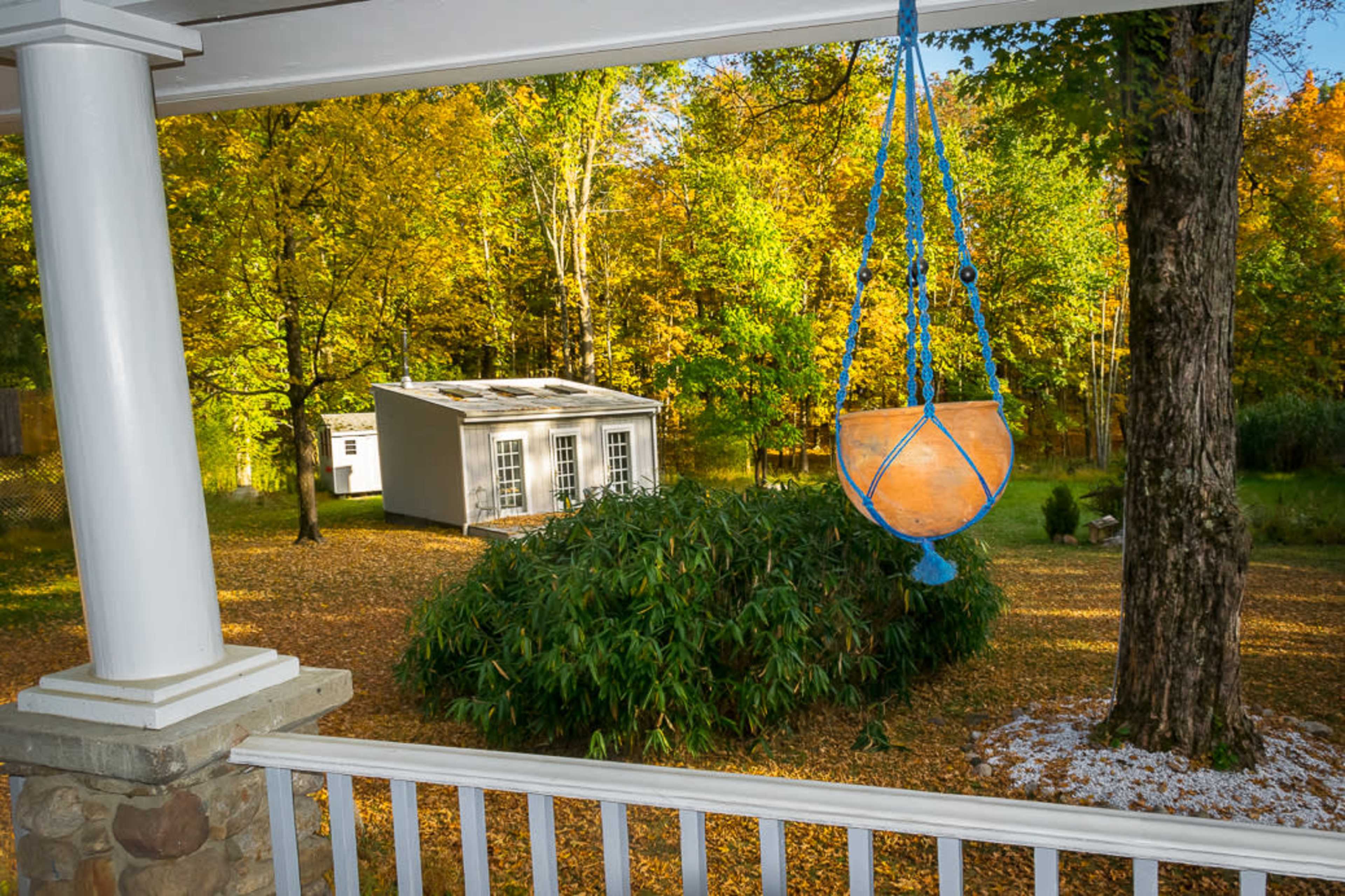 A swing hangs from a porch railing, overlooking a grassy area with a small white shed surrounded by autumn foliage.