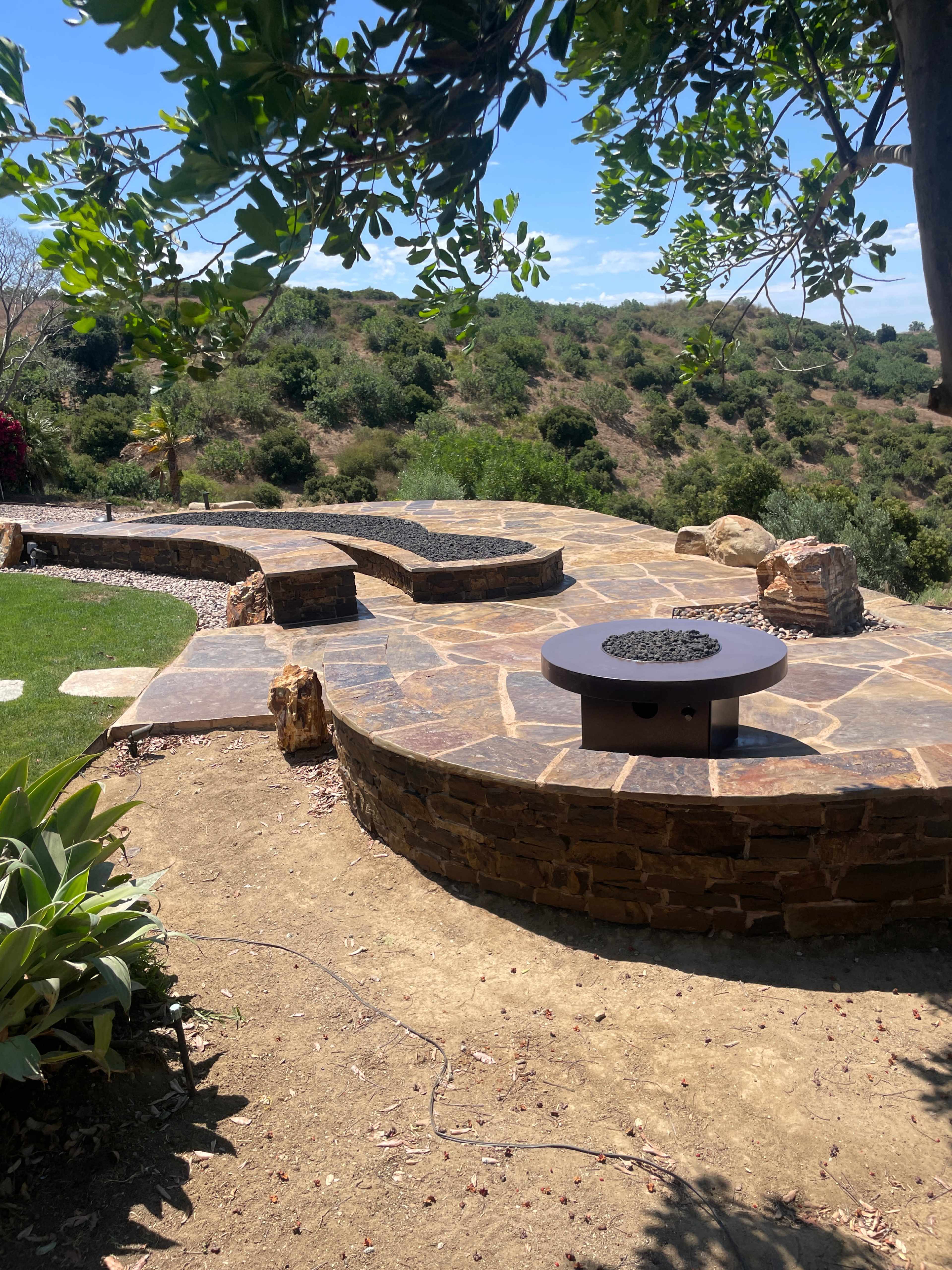 The image shows a landscaped outdoor area with a stone seating feature, surrounded by greenery and a fire pit at the center.