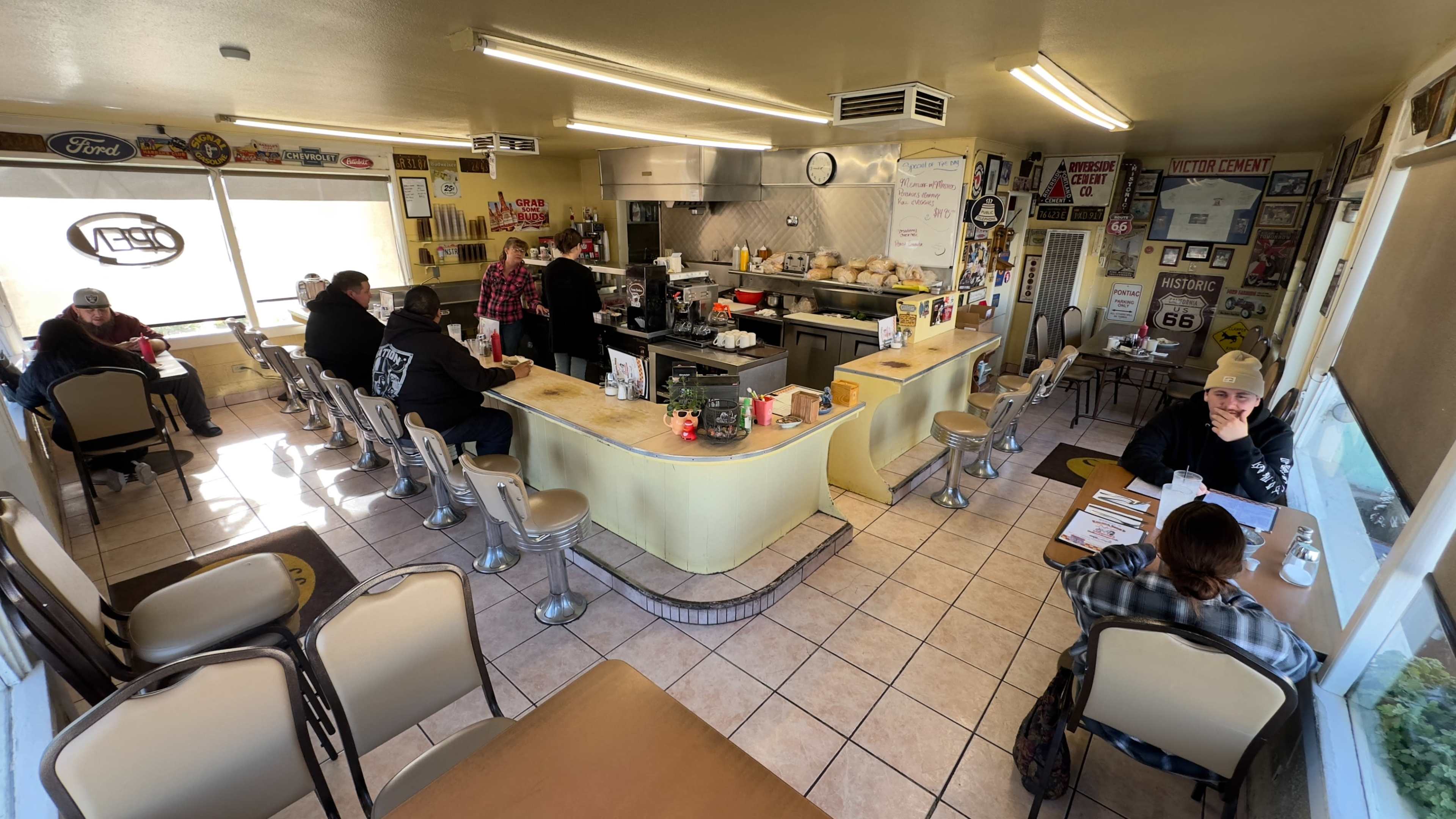 A diner with yellow walls and a counter serves customers seated at tables and chairs, while staff prepare food in the kitchen area.