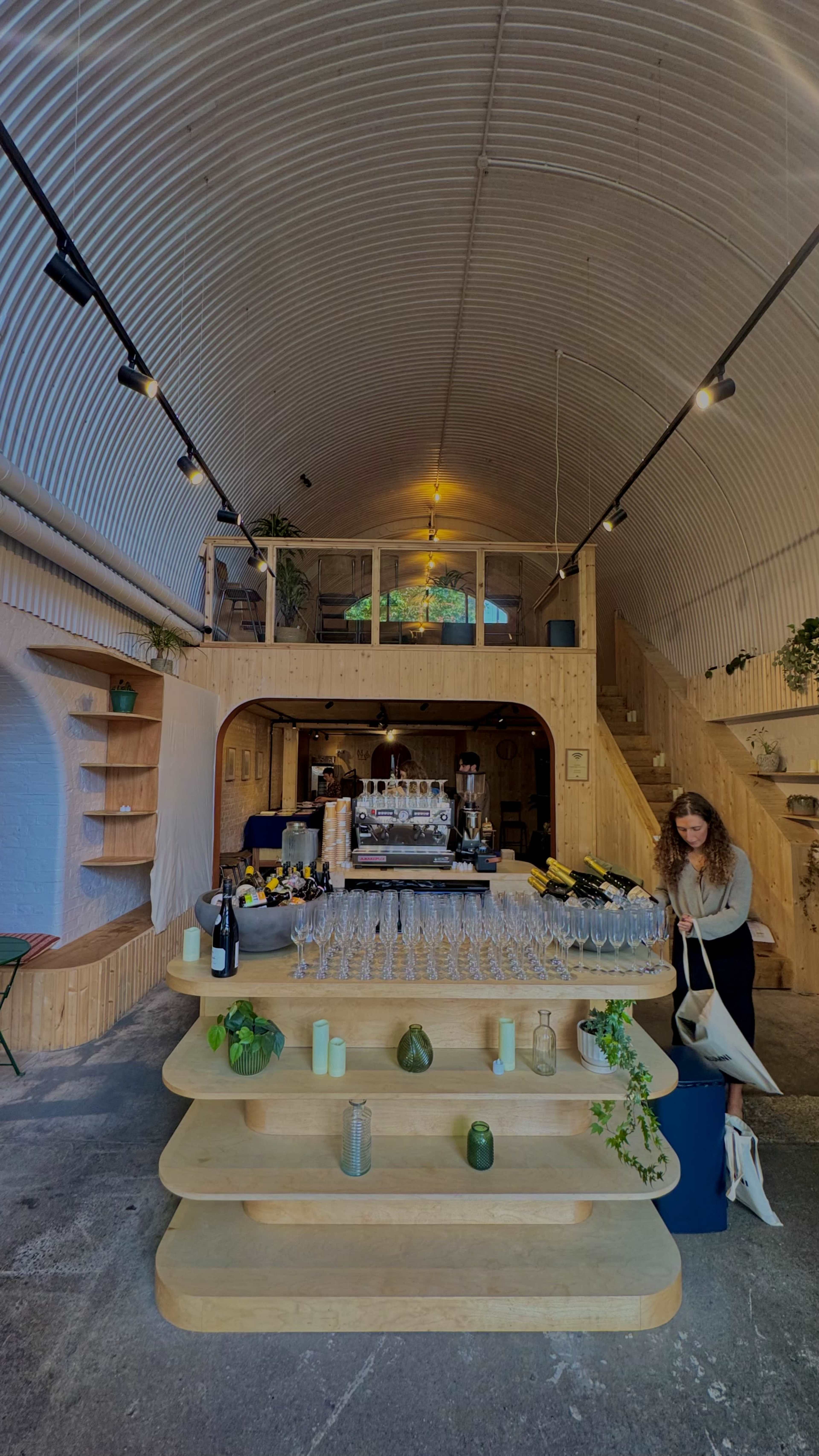 The image shows a modern wooden interior of a café or bar featuring a tiered display of glasses and decorative plants, with a woman arranging items in the foreground.
