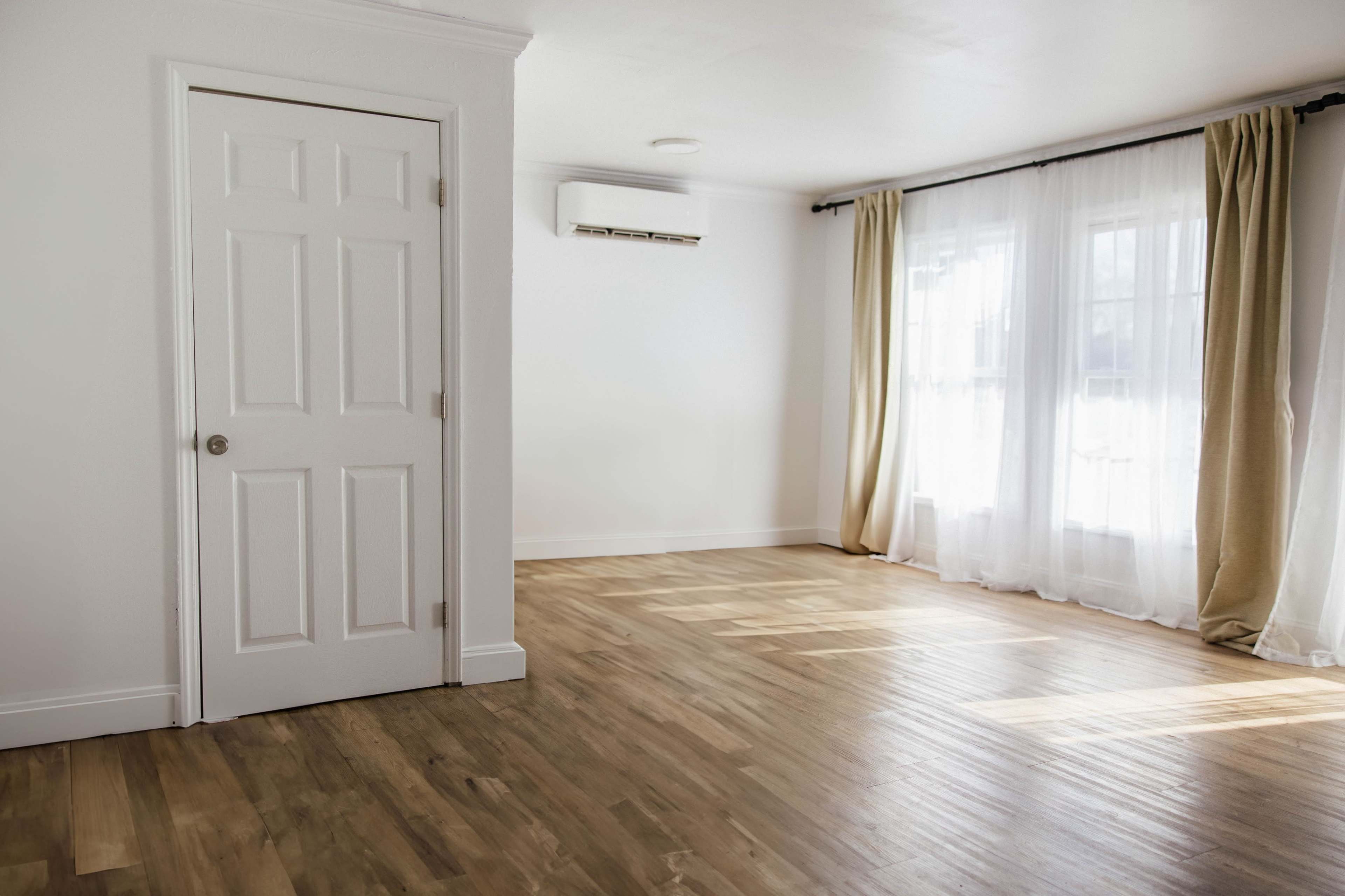 A bright, empty room with wooden flooring, a white door, and large windows covered by sheer curtains.