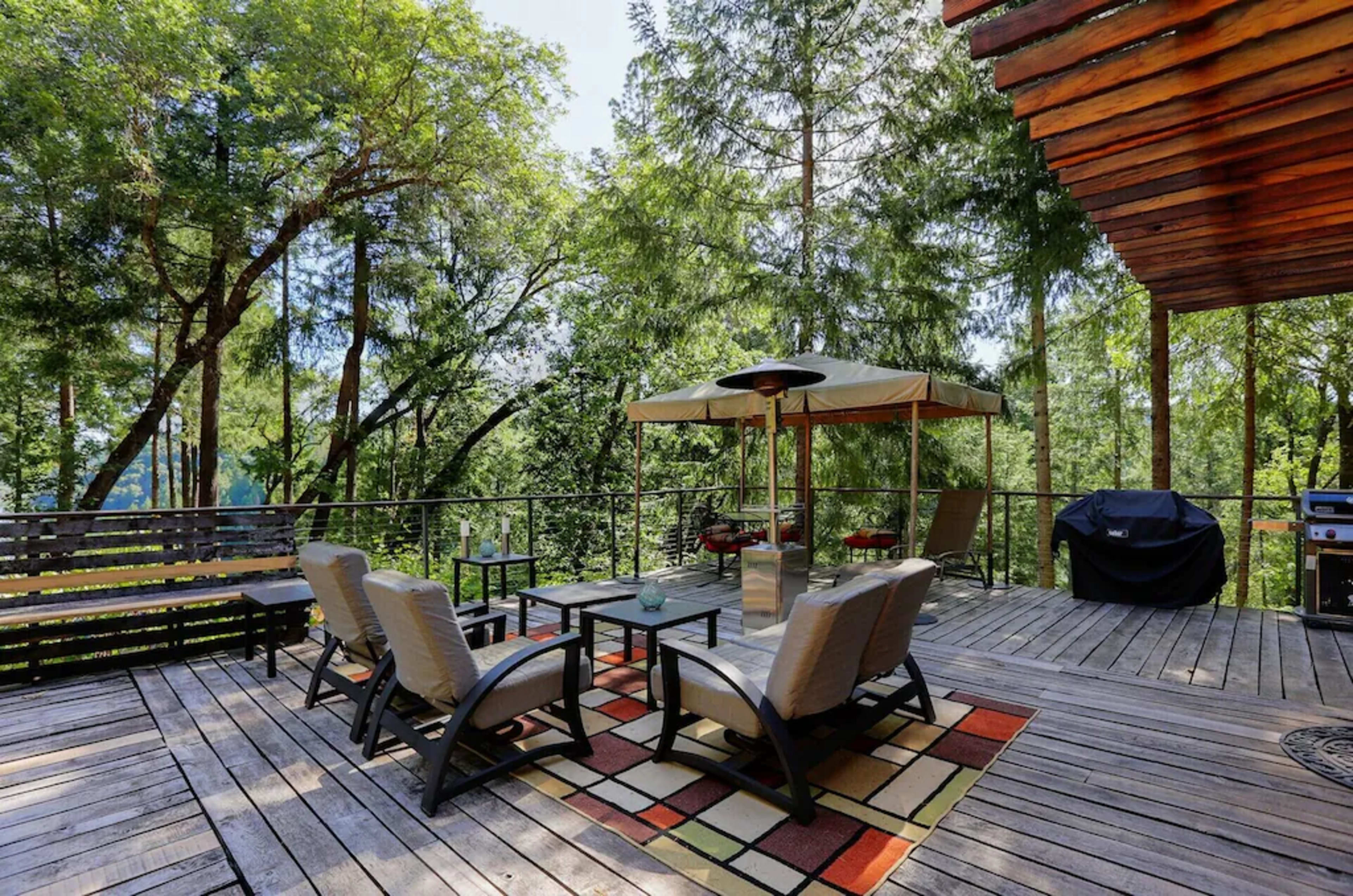 A wooden deck surrounded by trees, featuring seating arrangements, a canopy, and a grill.