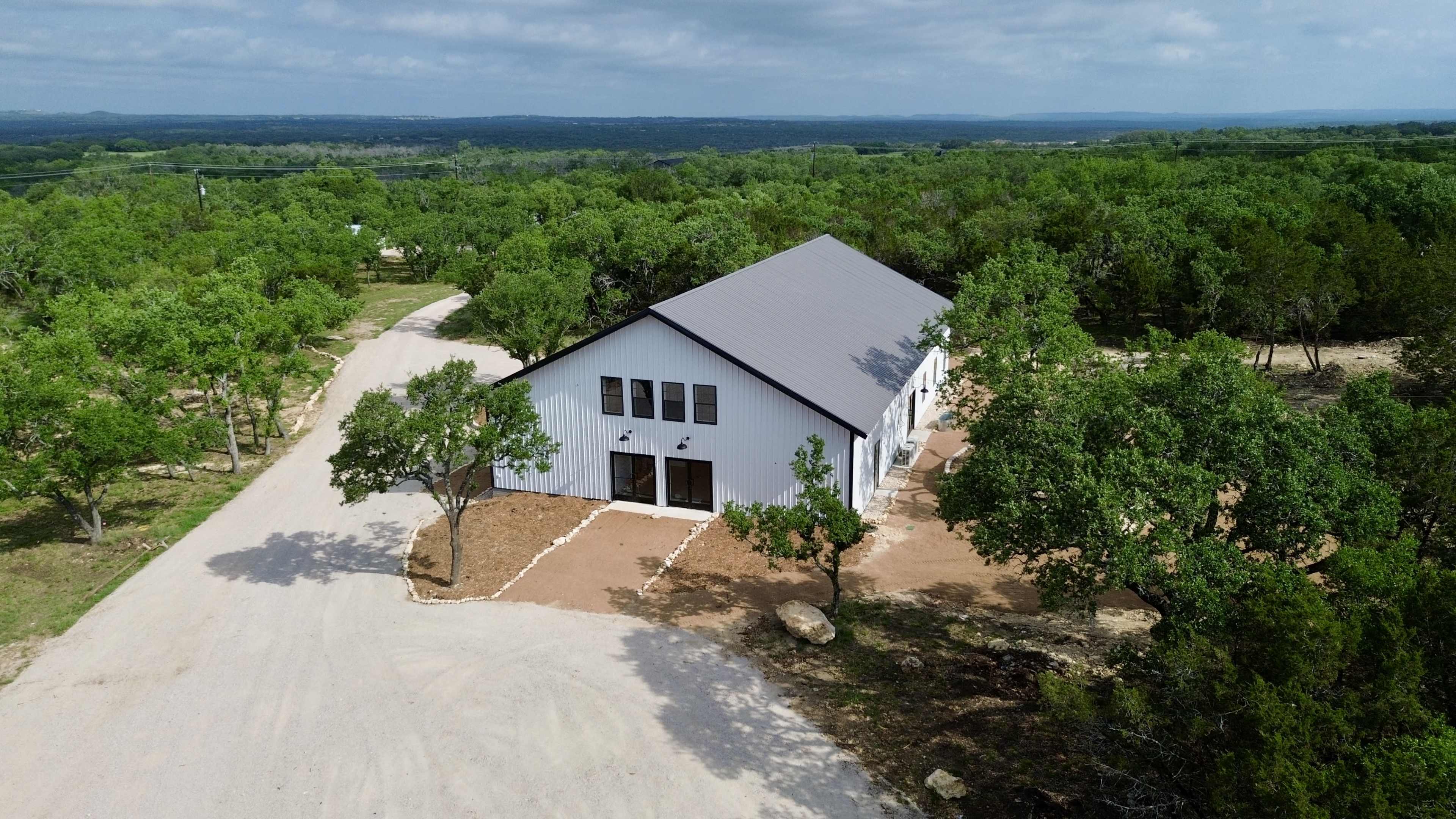 The image shows a large, modern building with a metal roof situated in a rural area surrounded by trees, with a dirt road leading up to it.