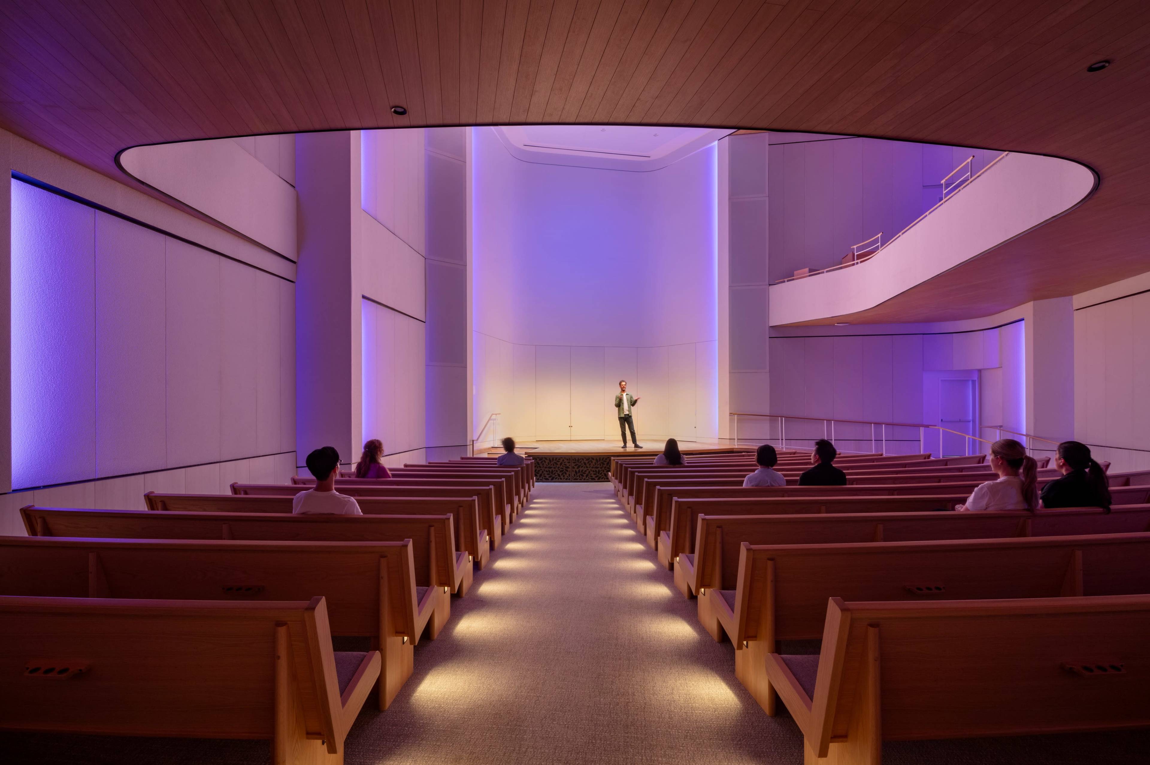 A speaker stands on a stage at the front of a modern, dimly lit auditorium, facing an audience seated on wooden benches.