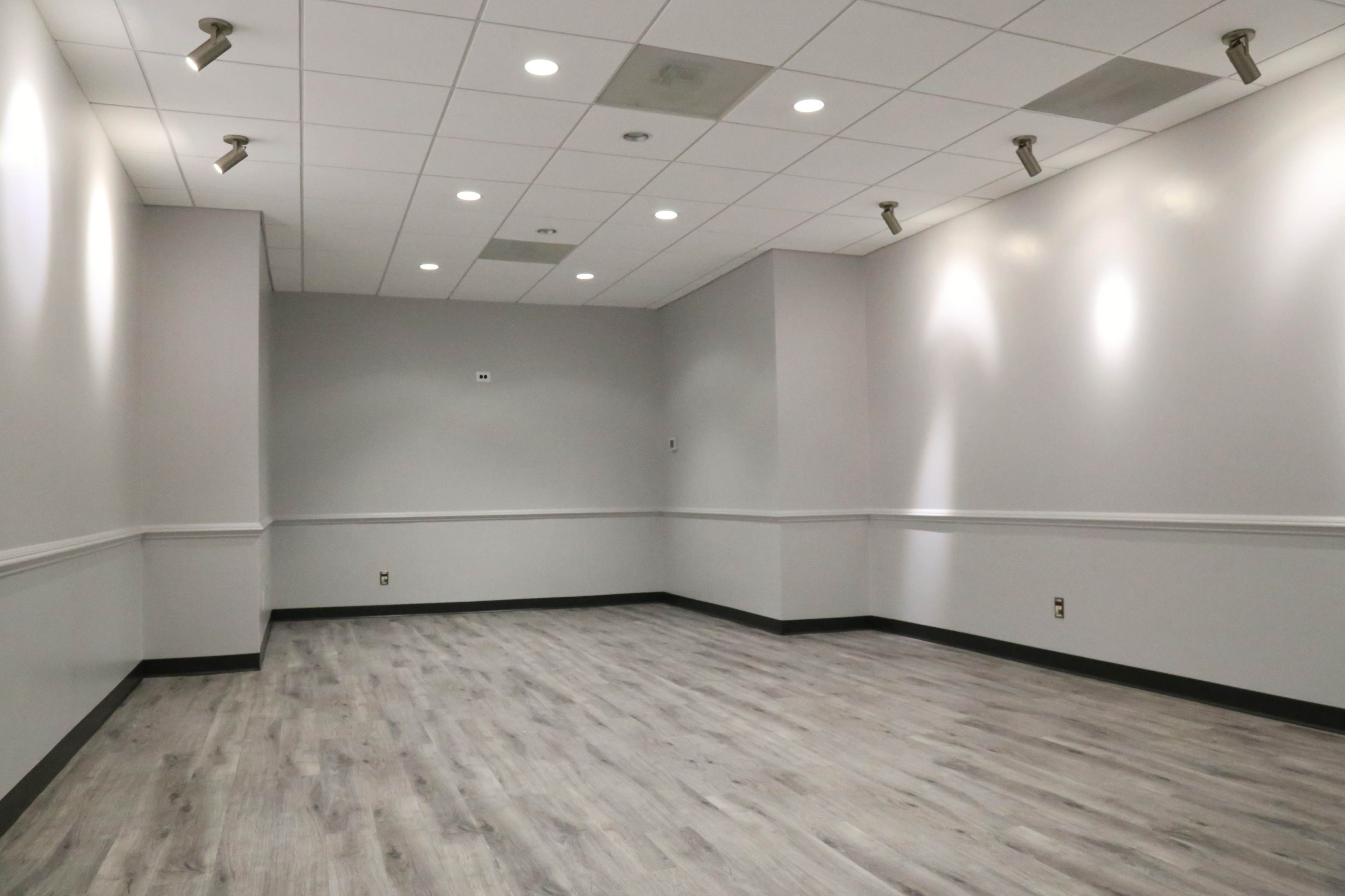 The image shows an empty, well-lit room with gray walls and a wooden floor, featuring track lighting on the ceiling.