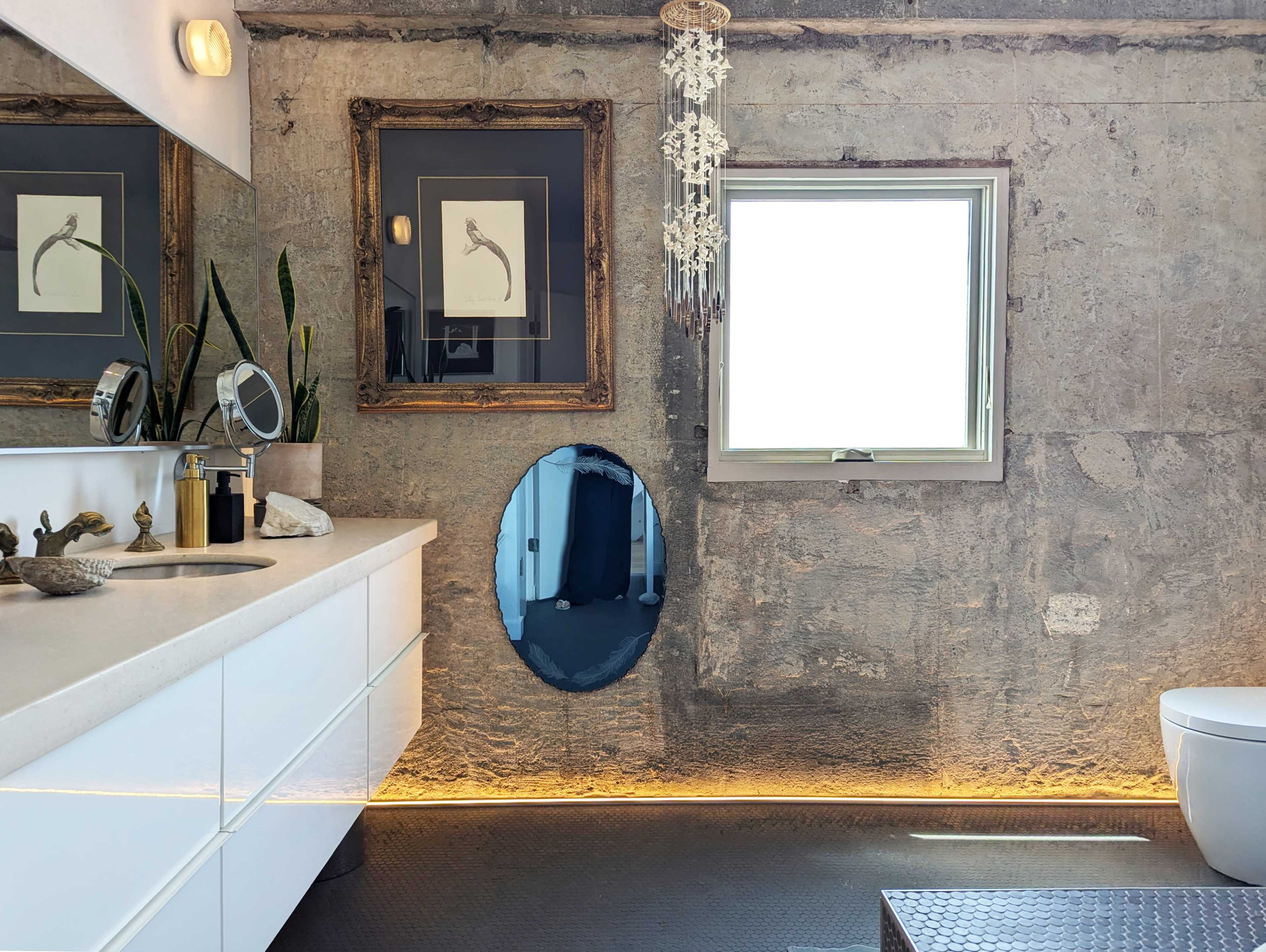 A modern bathroom features a minimalist vanity with a round mirror, distinct wall art, and natural stone walls illuminated by ambient lighting.