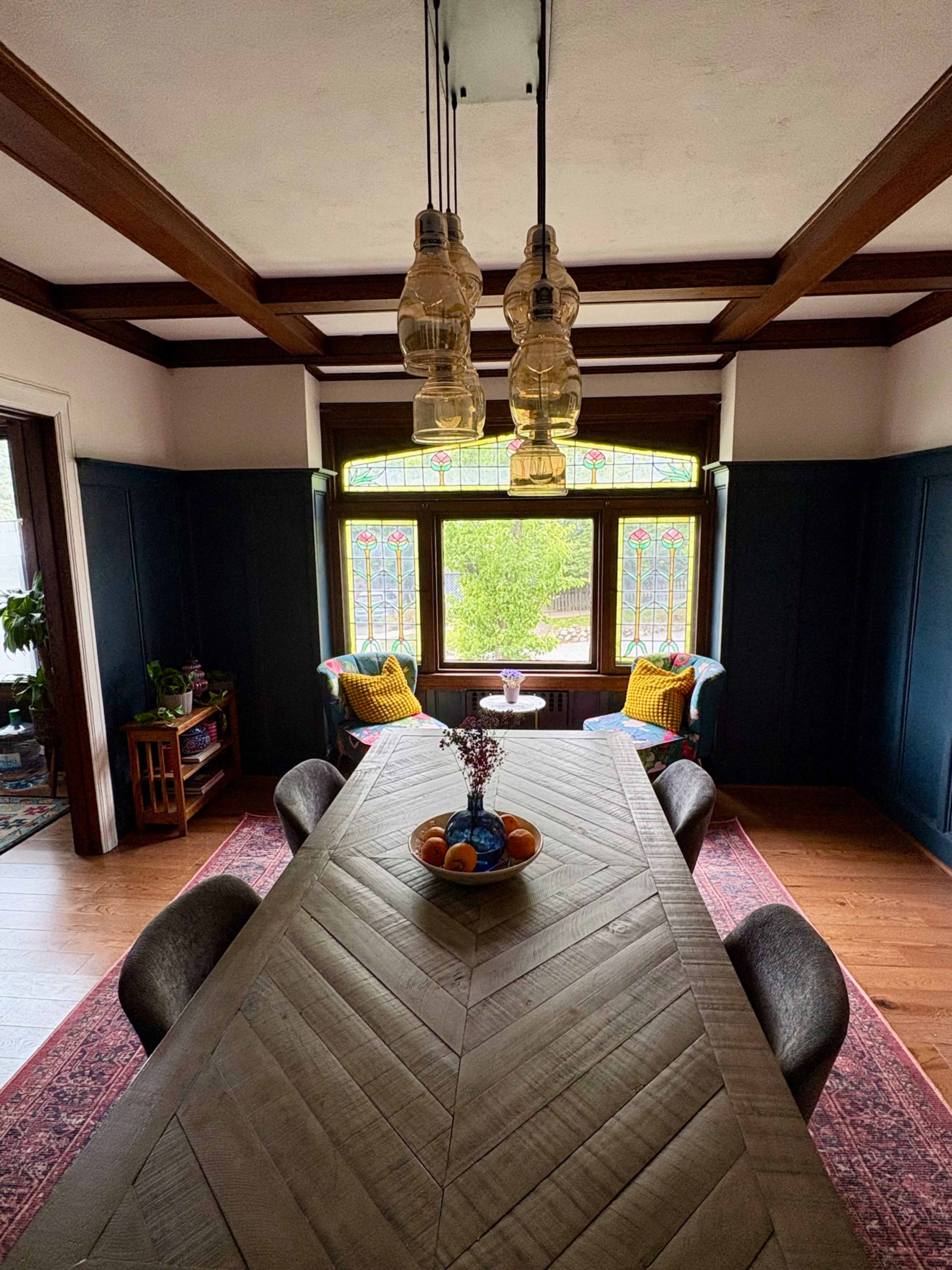 A long wooden dining table is set in a room with dark blue walls, featuring large windows that overlook a garden, and illuminated by three hanging glass lamps.