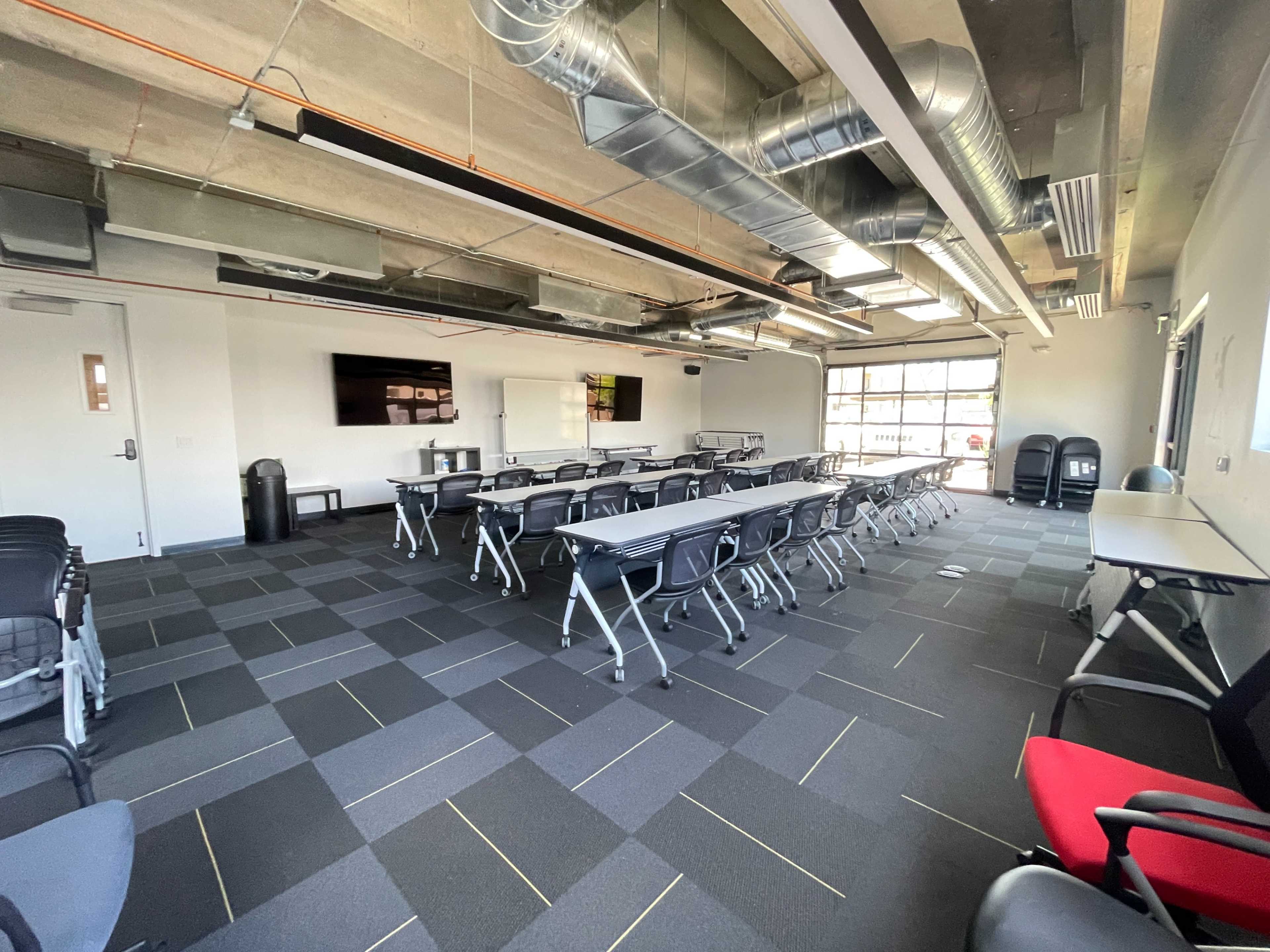 A spacious classroom with rows of tables and chairs, large windows, and a presentation screen on the wall.