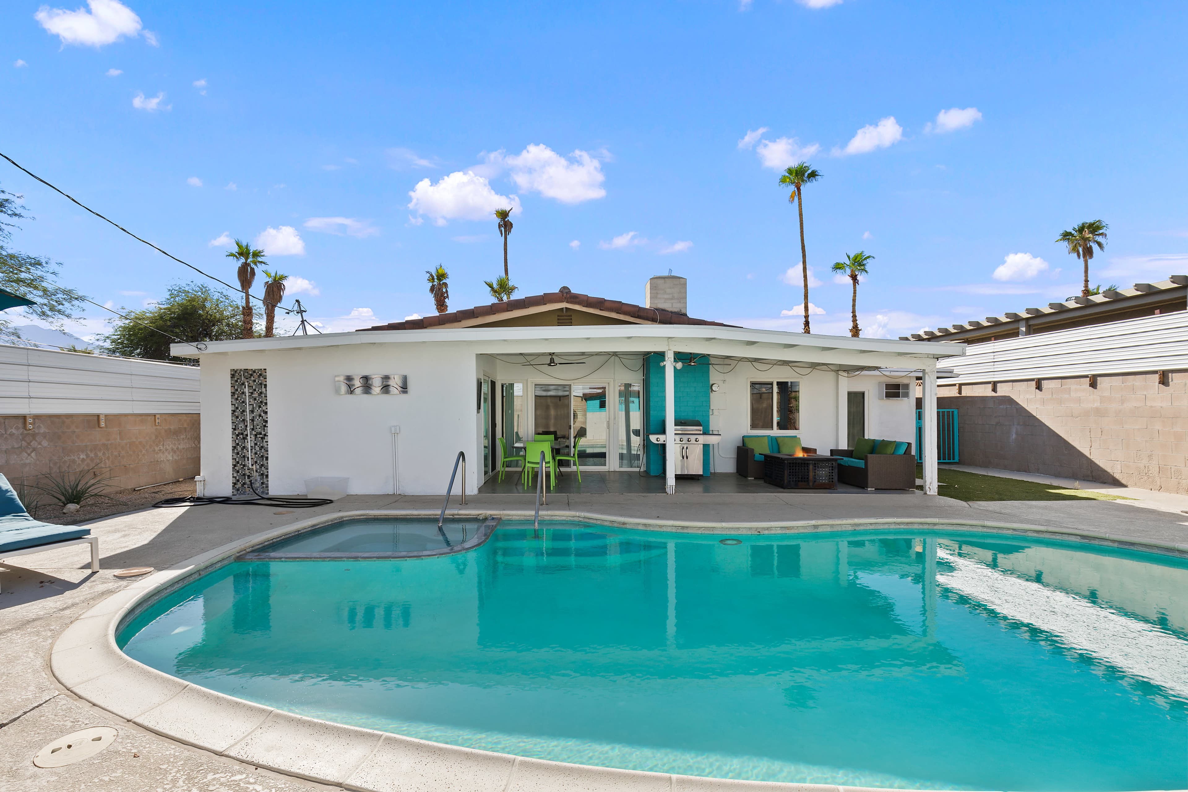 A residential backyard features a swimming pool next to a one-story house with palm trees in the background.