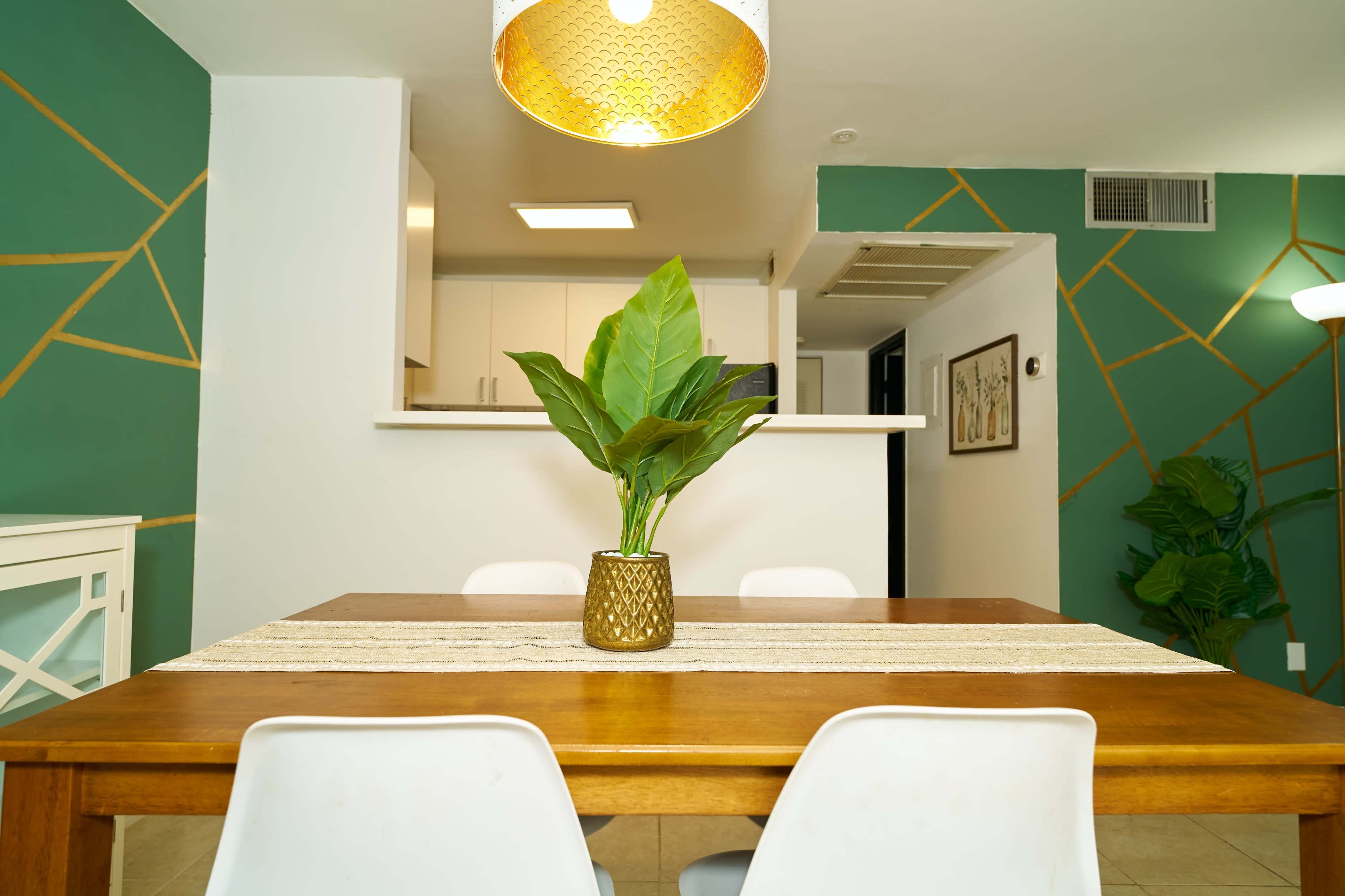 A dining table with a plant centerpiece is placed in a room with green walls featuring gold geometric patterns and a kitchen in the background.