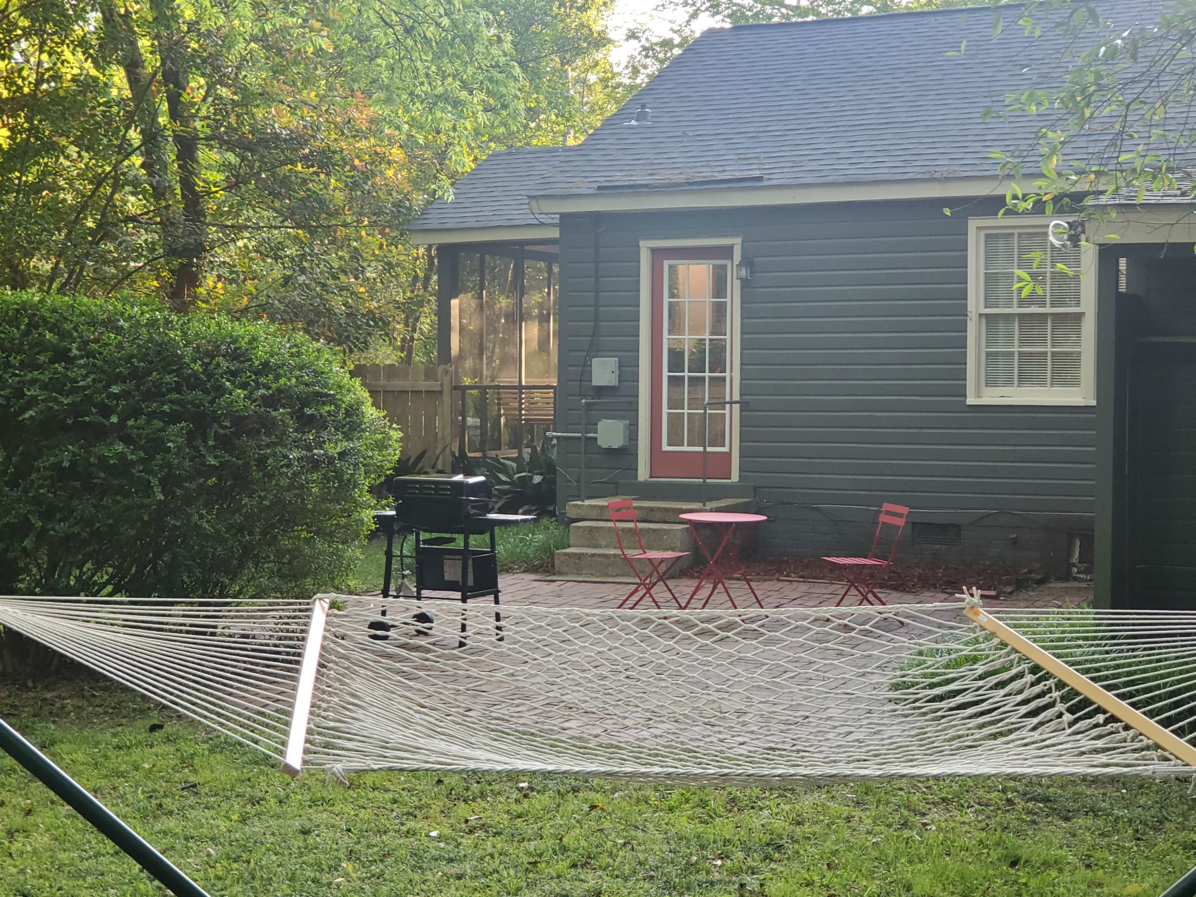 A hammock is strung between two supports in a yard with a grill and a small red table set in front of a green shrub and a gray house.