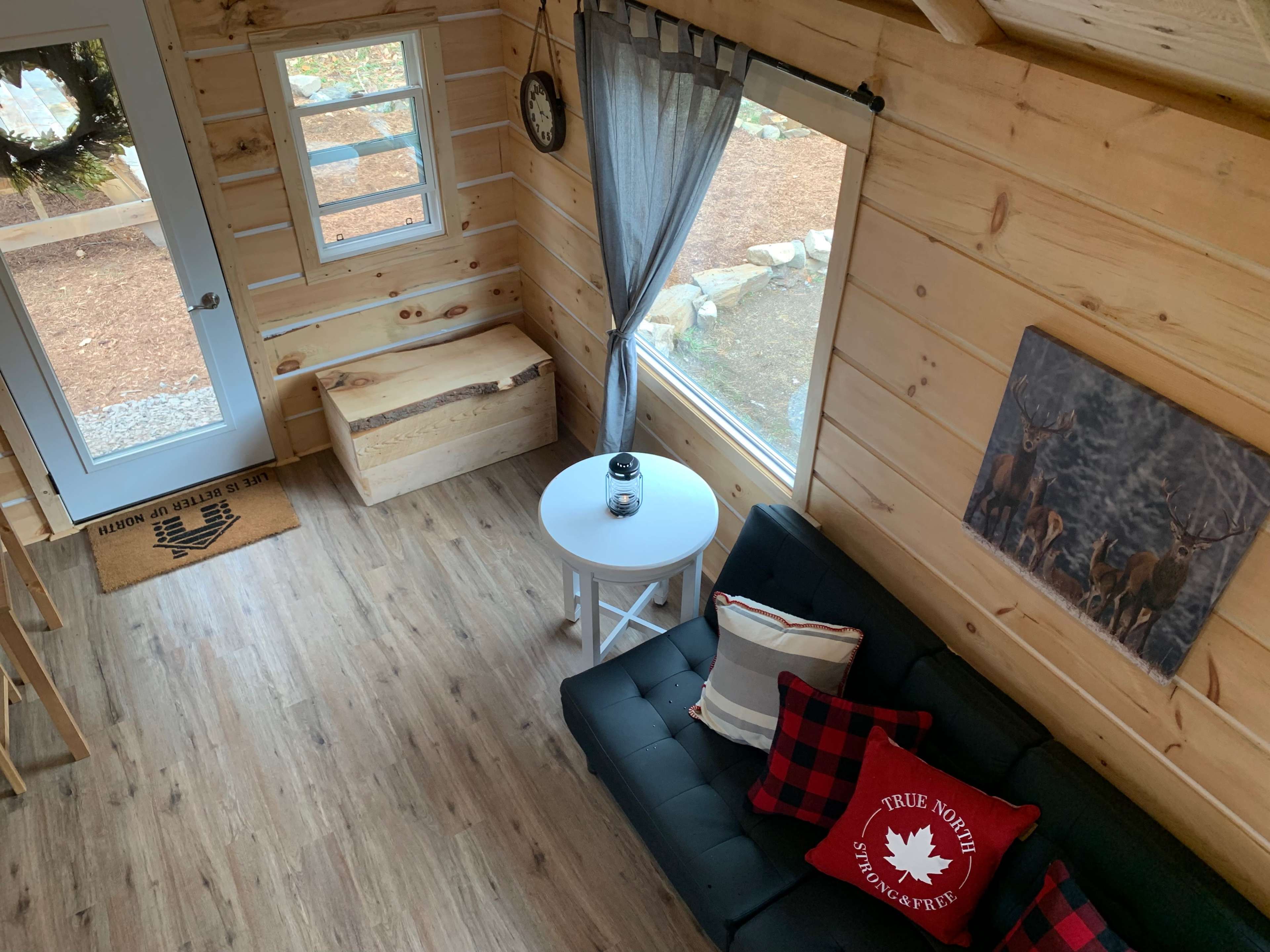 The image shows a small log cabin interior with a dark sofa, a round white table, and a window overlooking a gravel path outside.