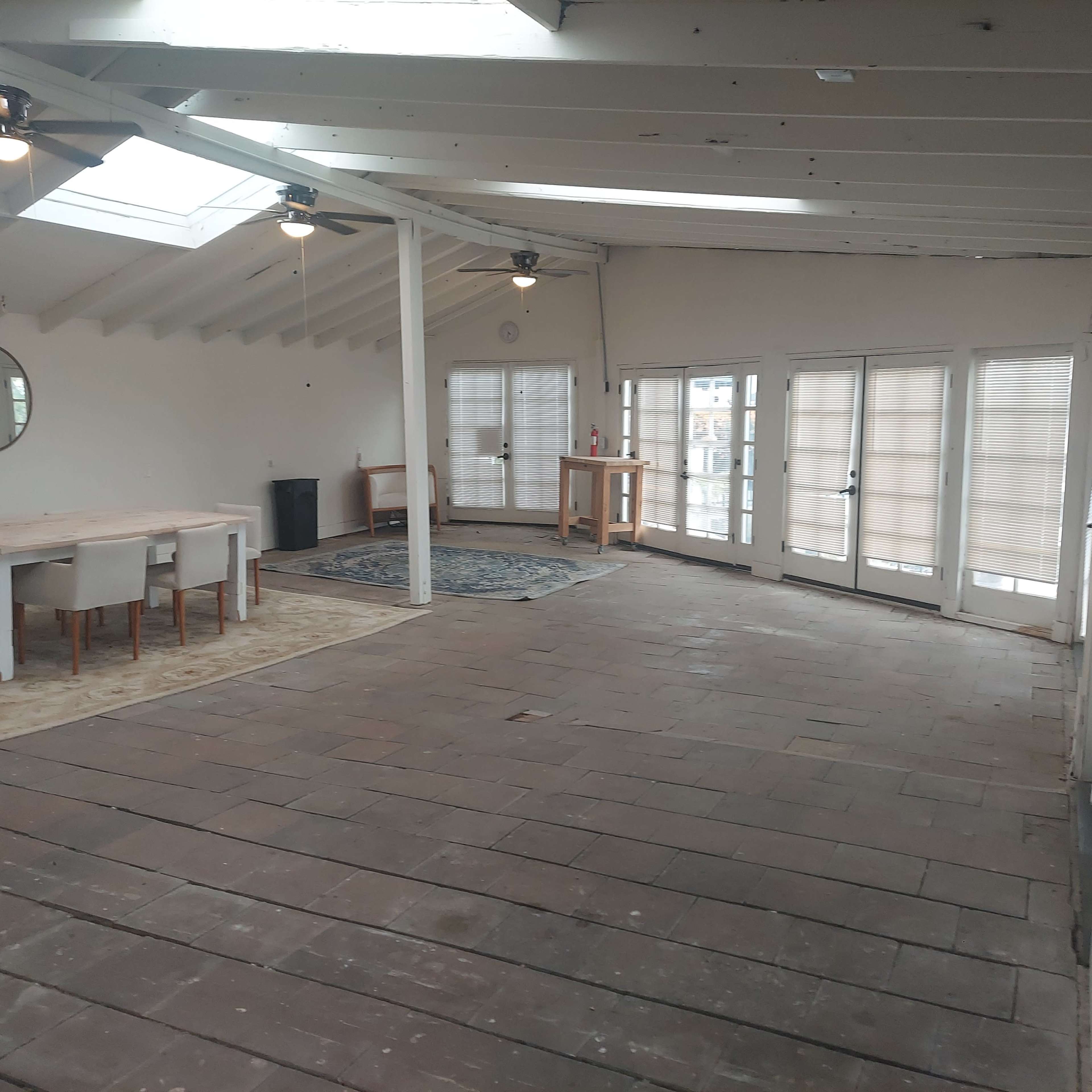 The image depicts an empty, open space with wooden flooring, a circular mirror on the wall, a dining table with chairs, and large windows leading to an outdoor area.