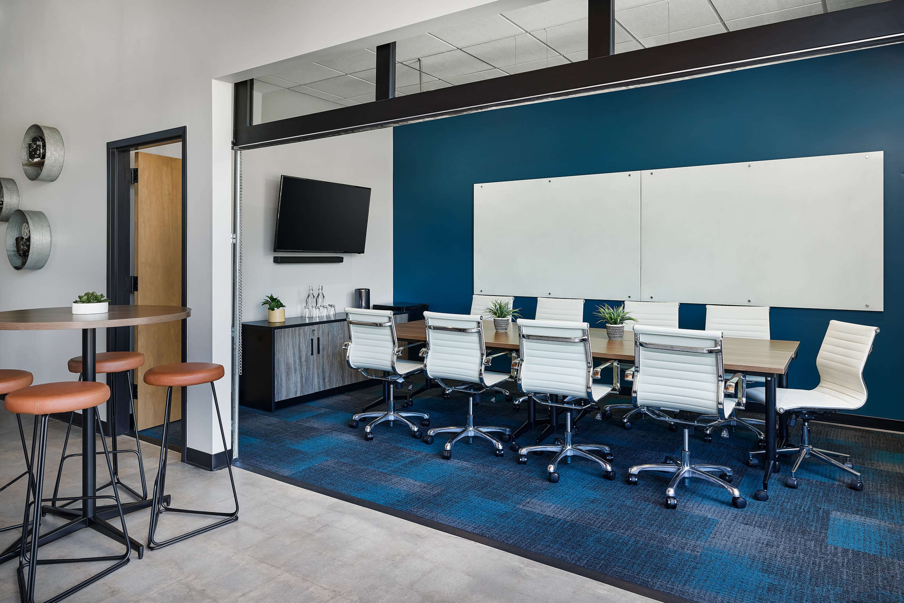 A modern conference room with a rectangular table surrounded by white swivel chairs, a television mounted on the wall, and a large whiteboard.