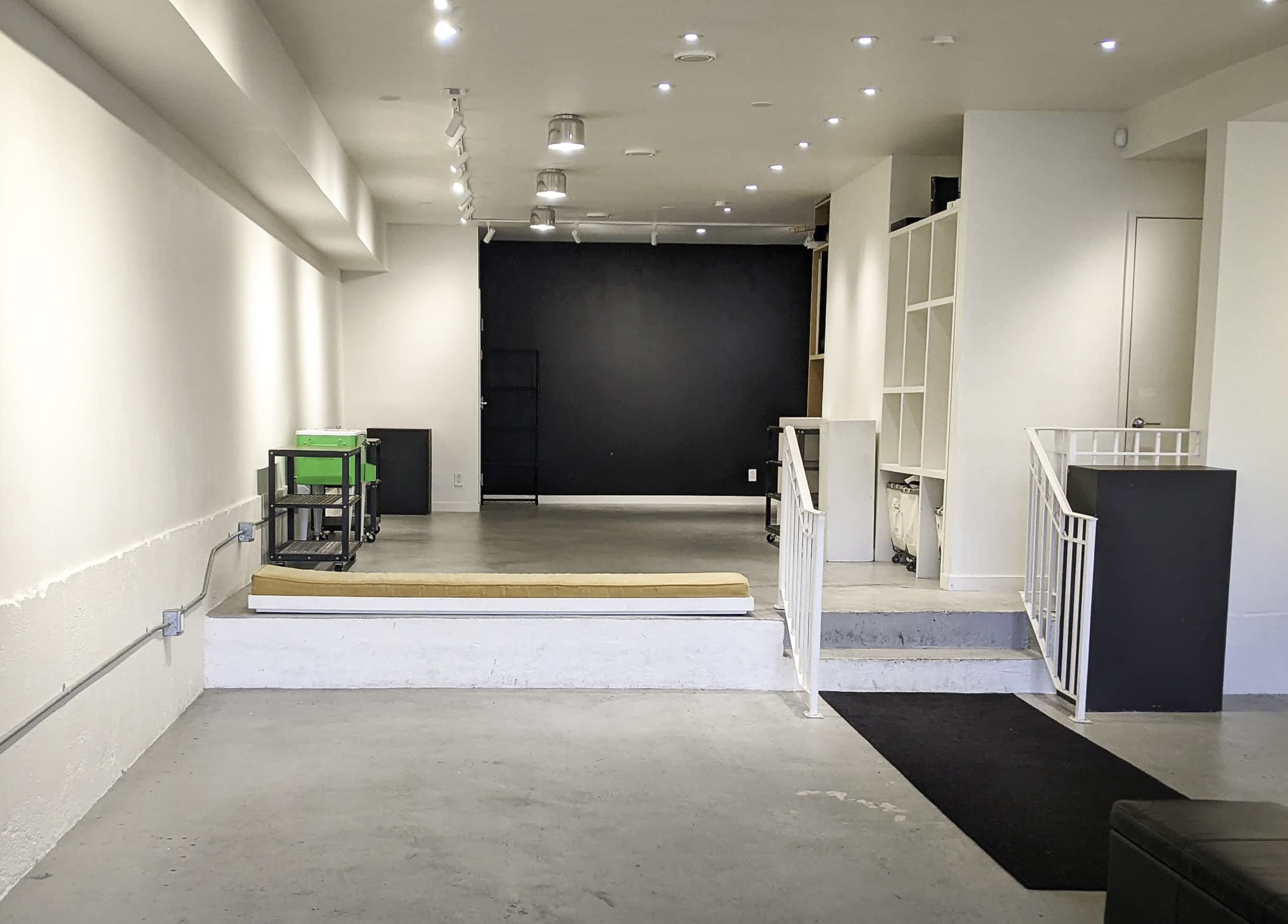 A minimalist interior space featuring a black wall, a raised platform with a mattress, a few scattered furniture items, and overhead lighting.
