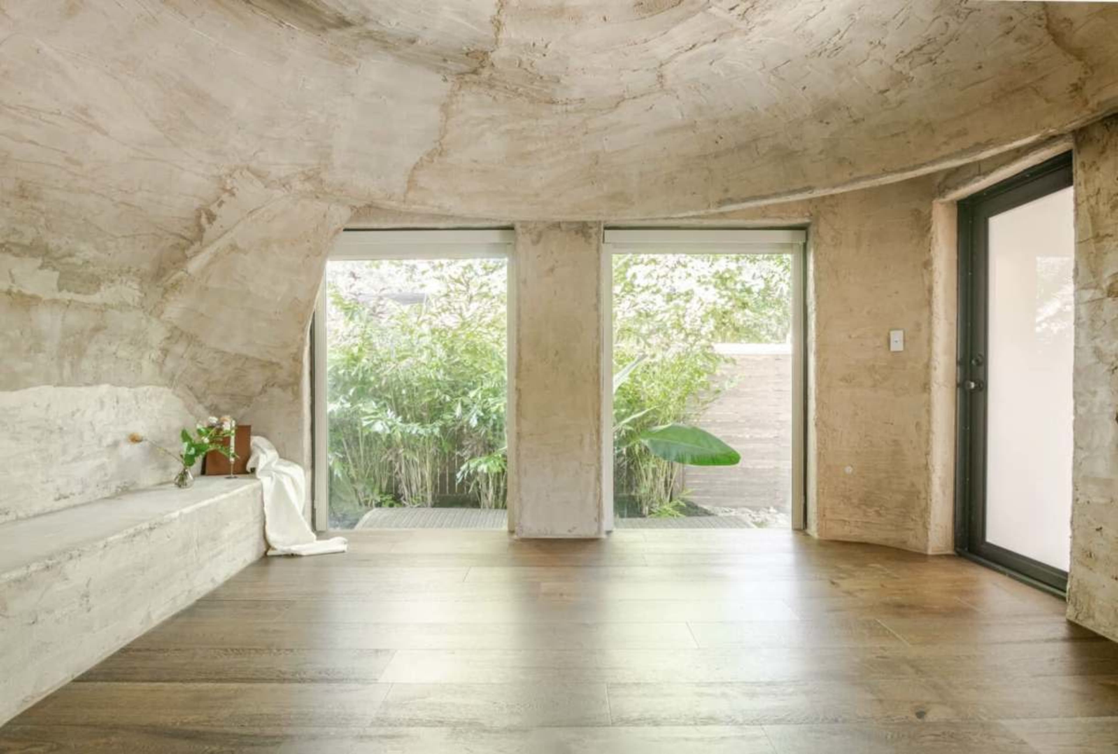 The image shows a minimalist, curved interior space with concrete walls, wooden flooring, and large glass doors leading to a garden.