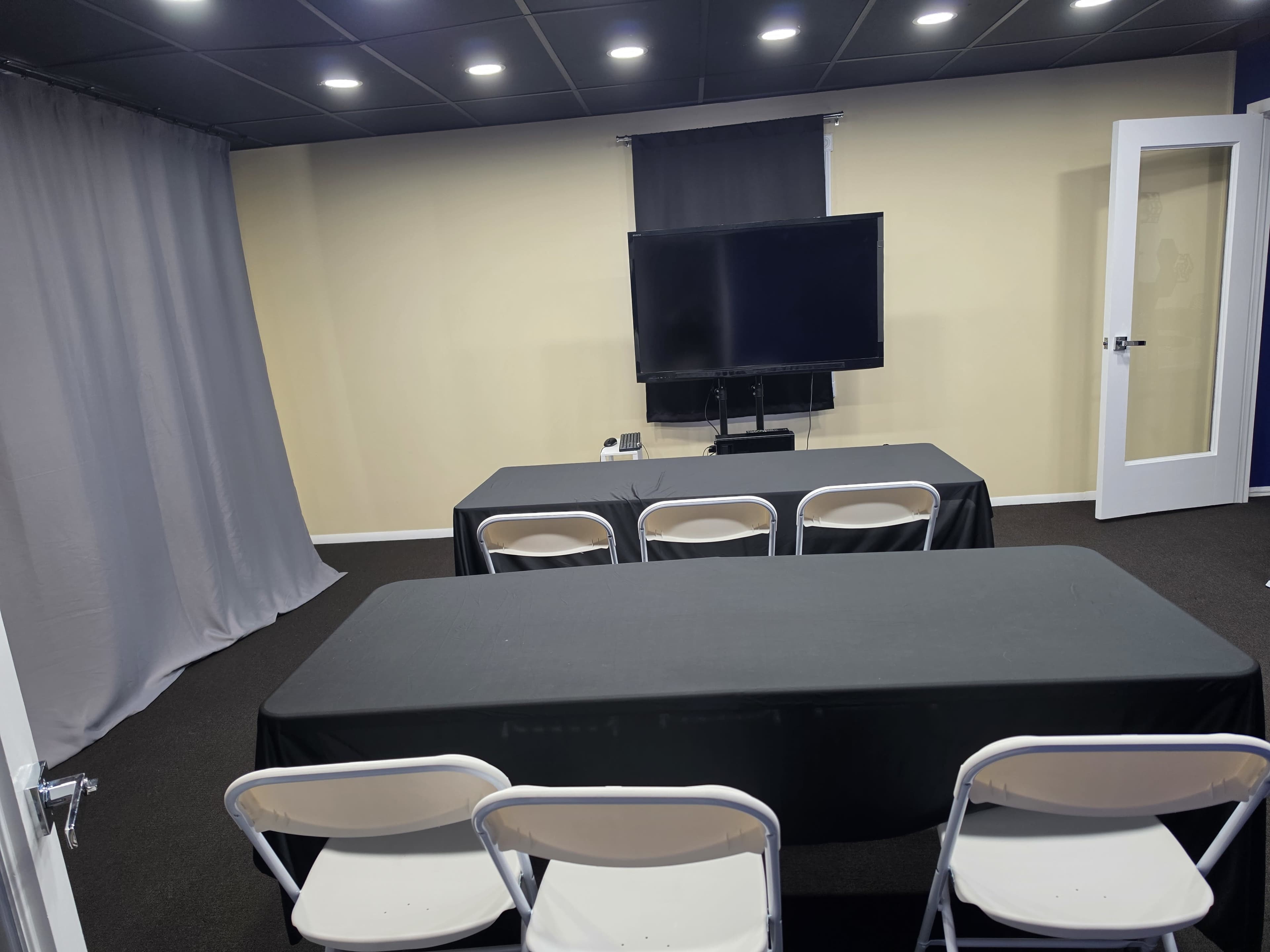 The image shows a small, prepared meeting room with two tables covered in black cloth, several white chairs, and a large television mounted on the wall.