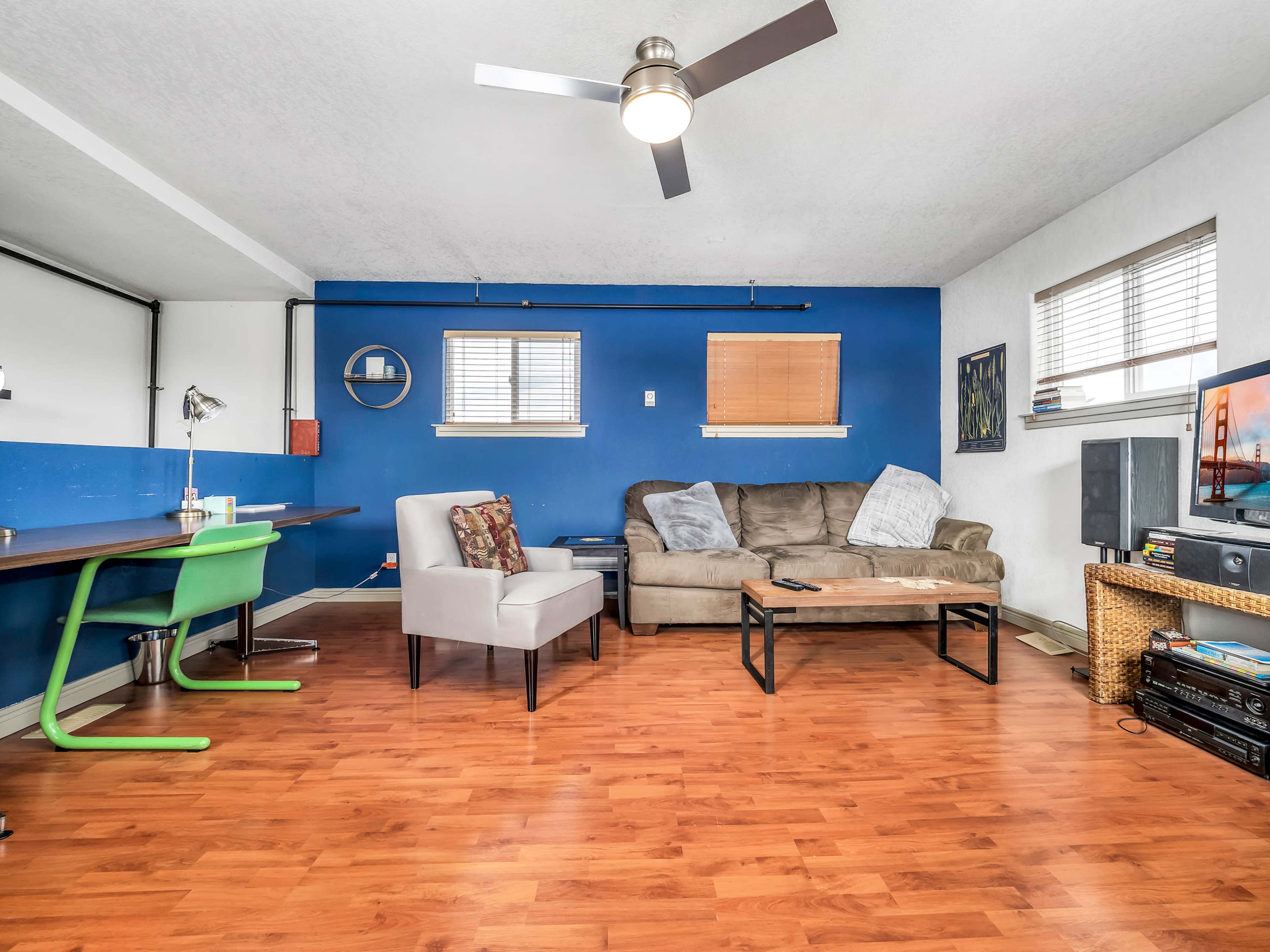 A living room with a blue accent wall, a brown couch, a wooden coffee table, a desk with a green chair, and a television.