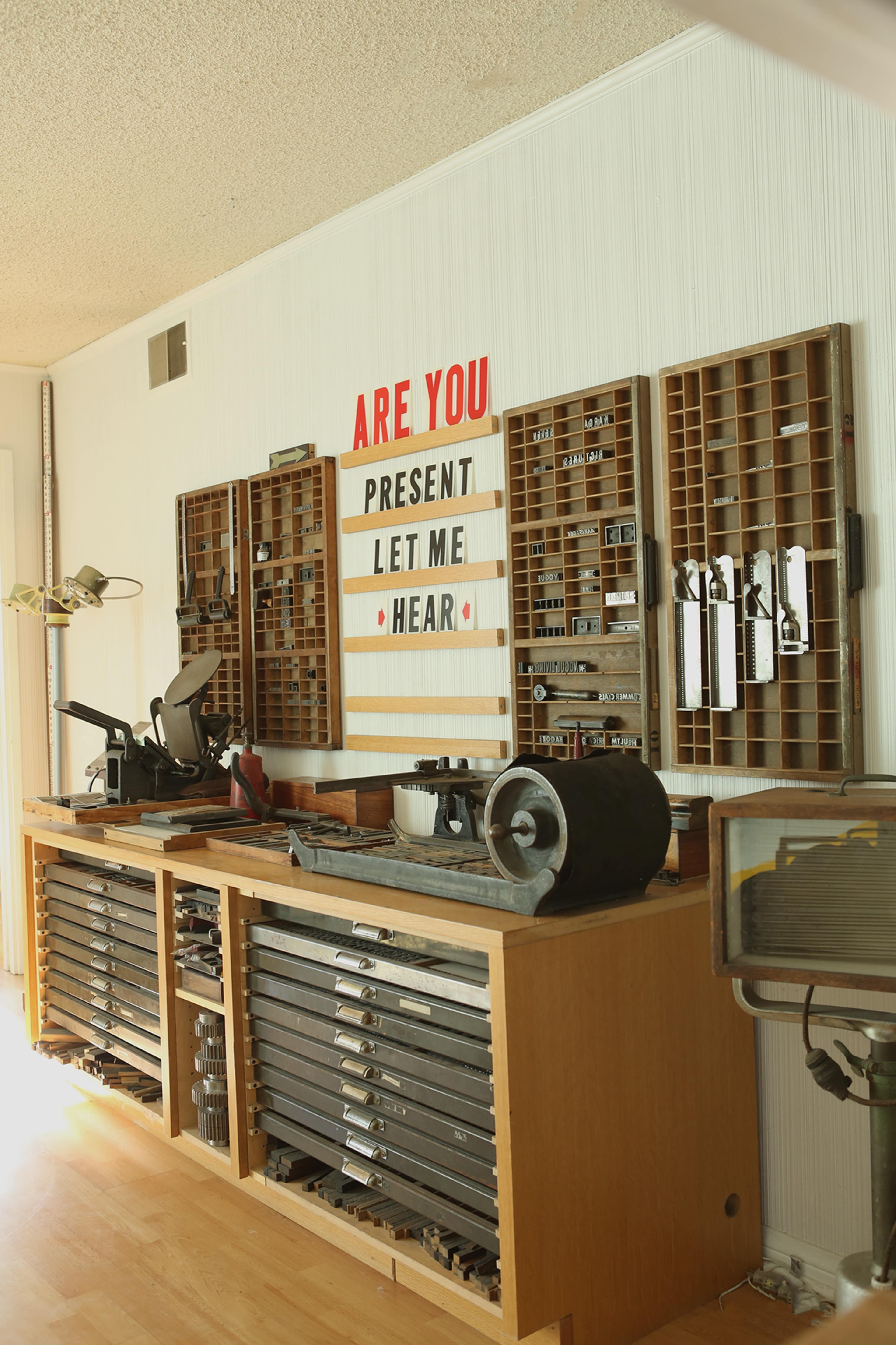 The image shows a woodworking workshop with organized tools, a large workbench, and a prominently displayed sign that reads, "ARE YOU PRESENT LET ME HEAR."